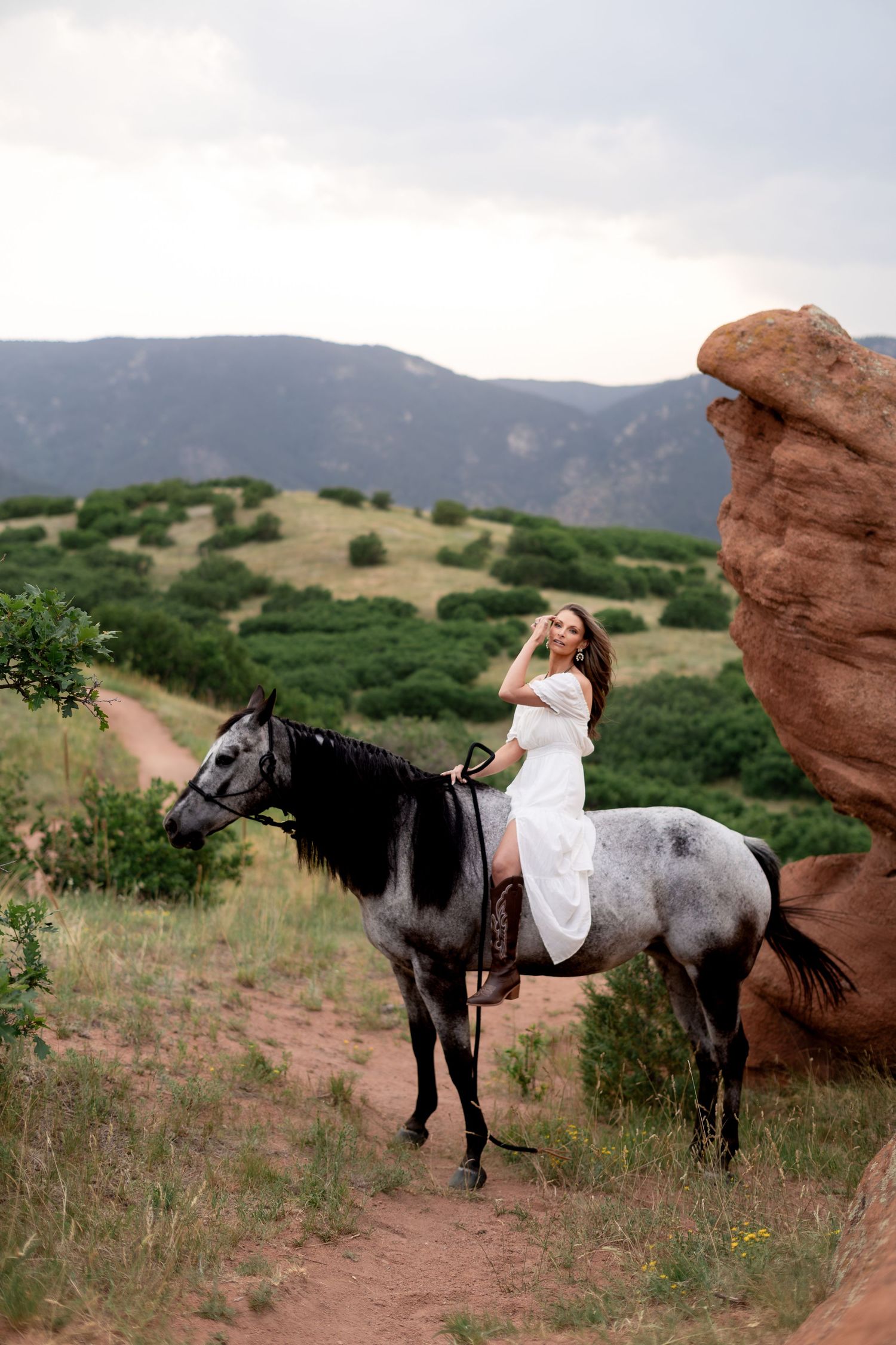 Woman in white dress riding a gray horse near red rock formations with green mountain landscape backdrop.