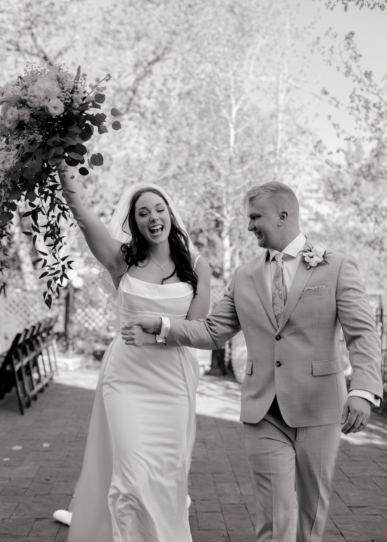 Joyful bride raising bouquet while walking with groom in gray suit after wedding ceremony.