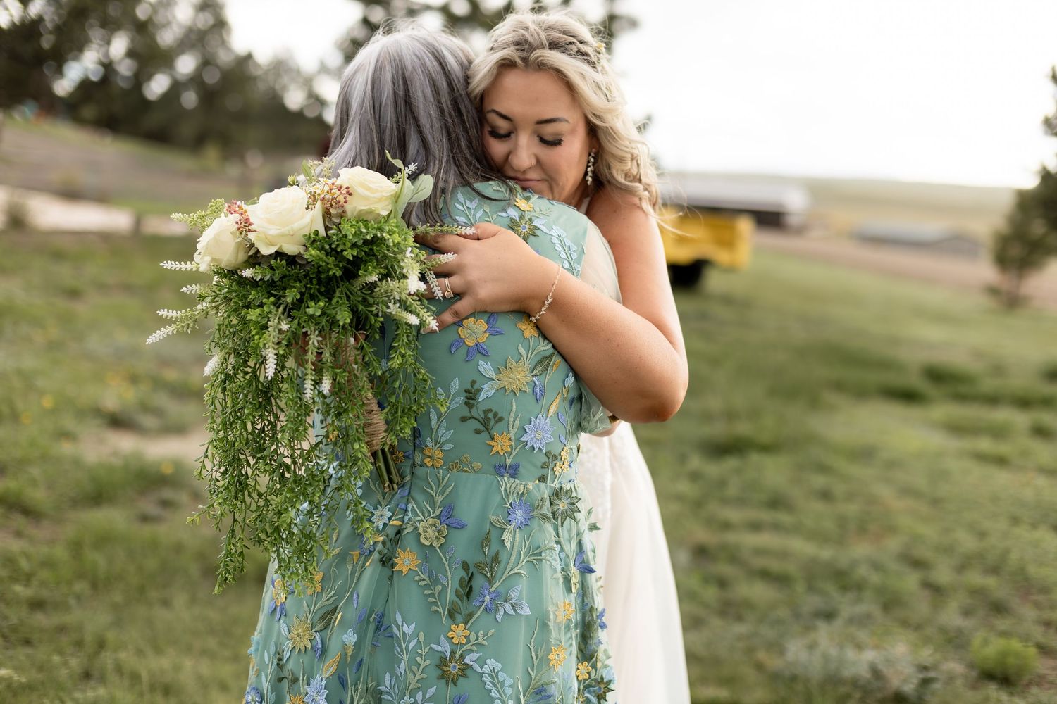 Bride in white gown embracing older woman holding white bouquet at outdoor wedding.