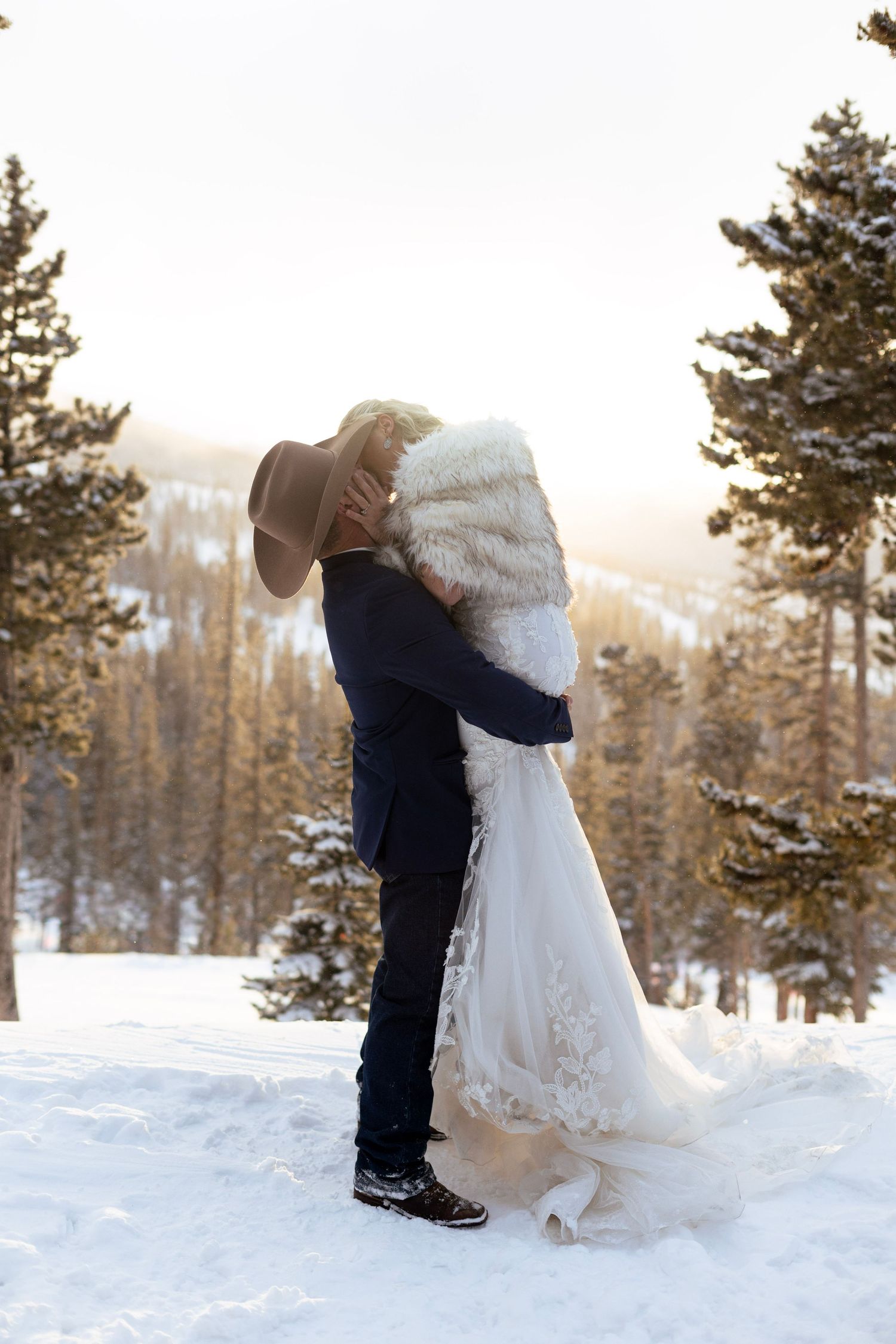 A couple shares a romantic winter wedding moment in the snow with evergreen trees and mountains in the background.