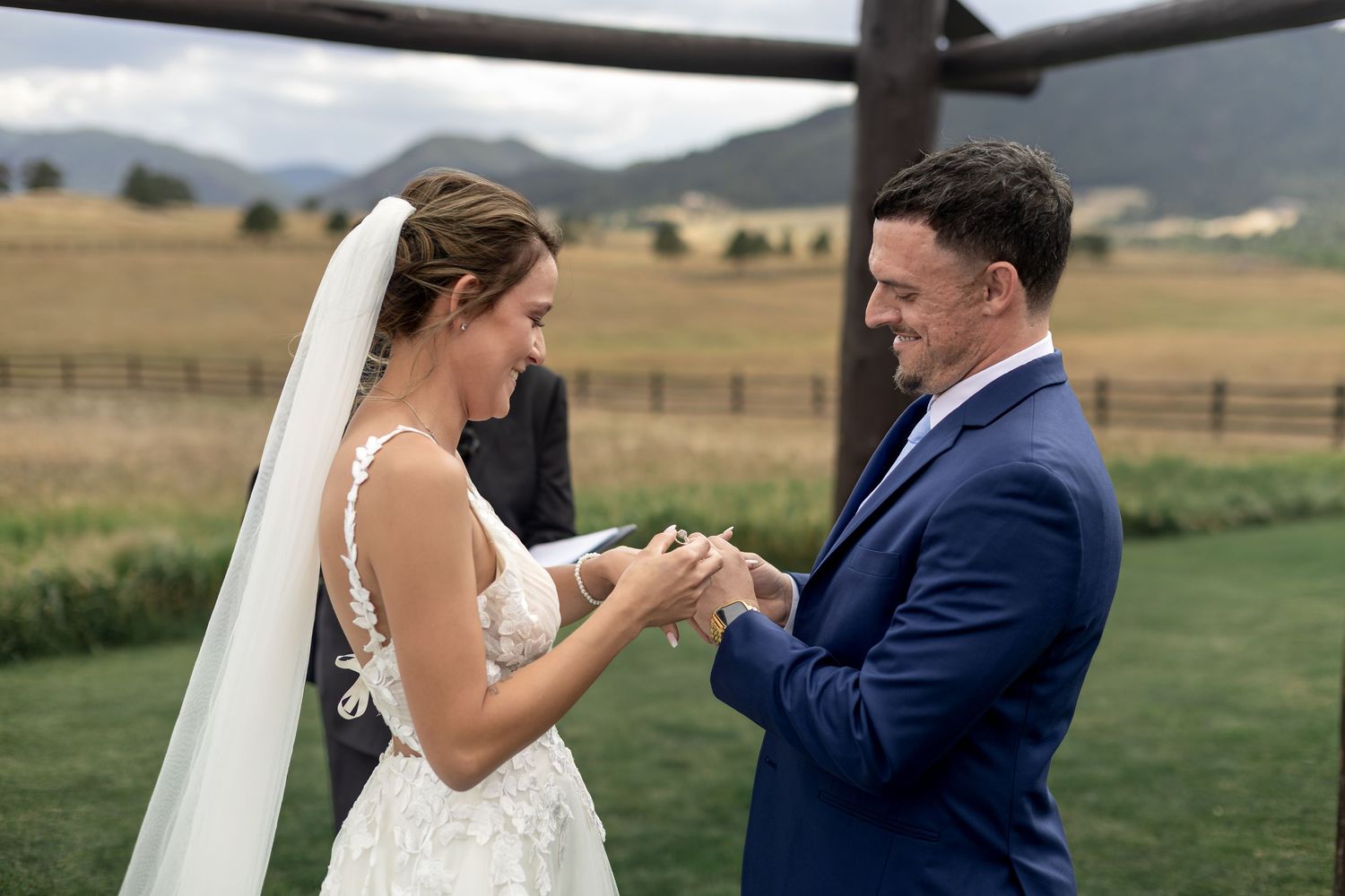 Bride placing wedding ring on groom's finger during outdoor ceremony with mountain backdrop.