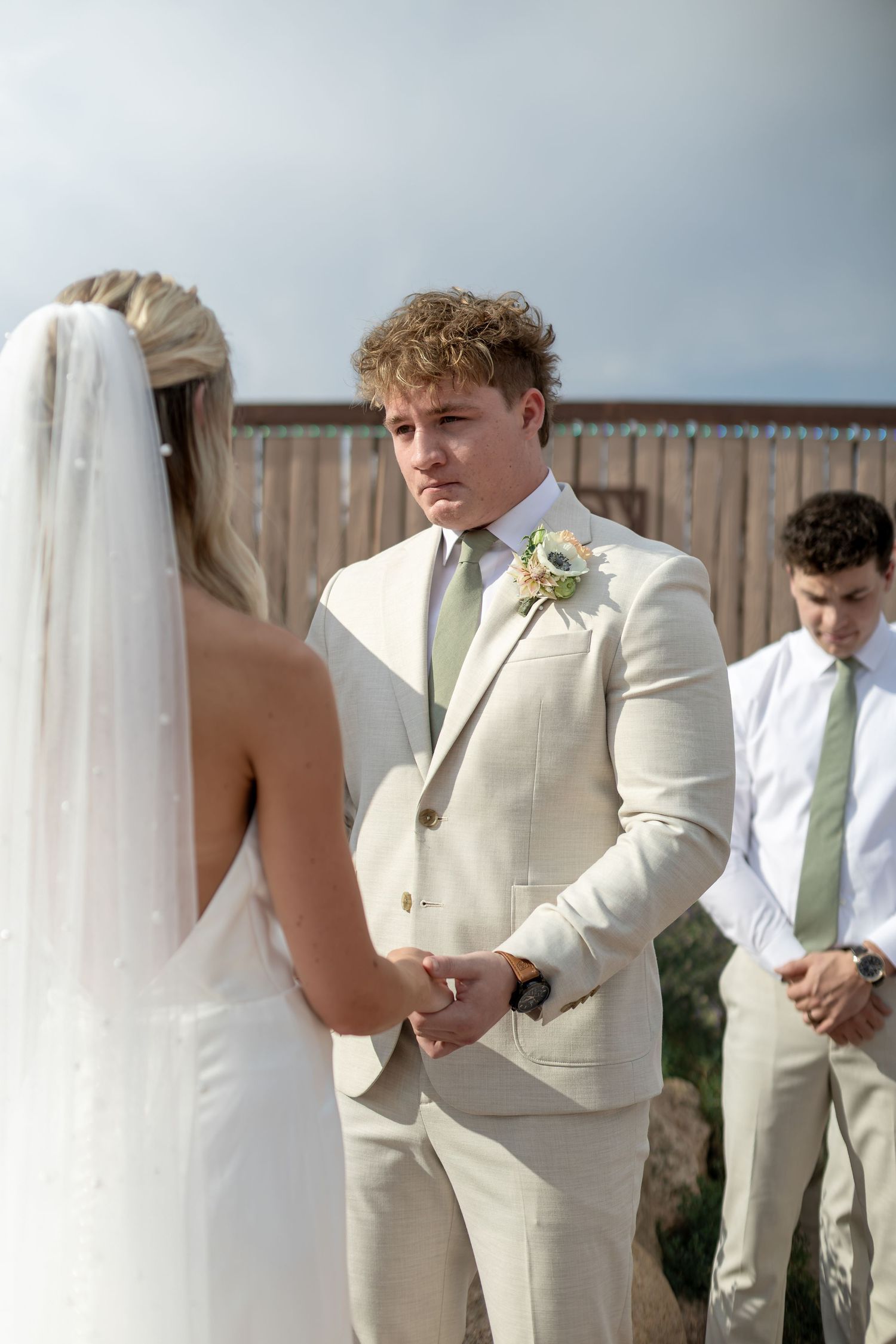 Groom in cream suit holds brides hands during outdoor wedding ceremony with guests in background.