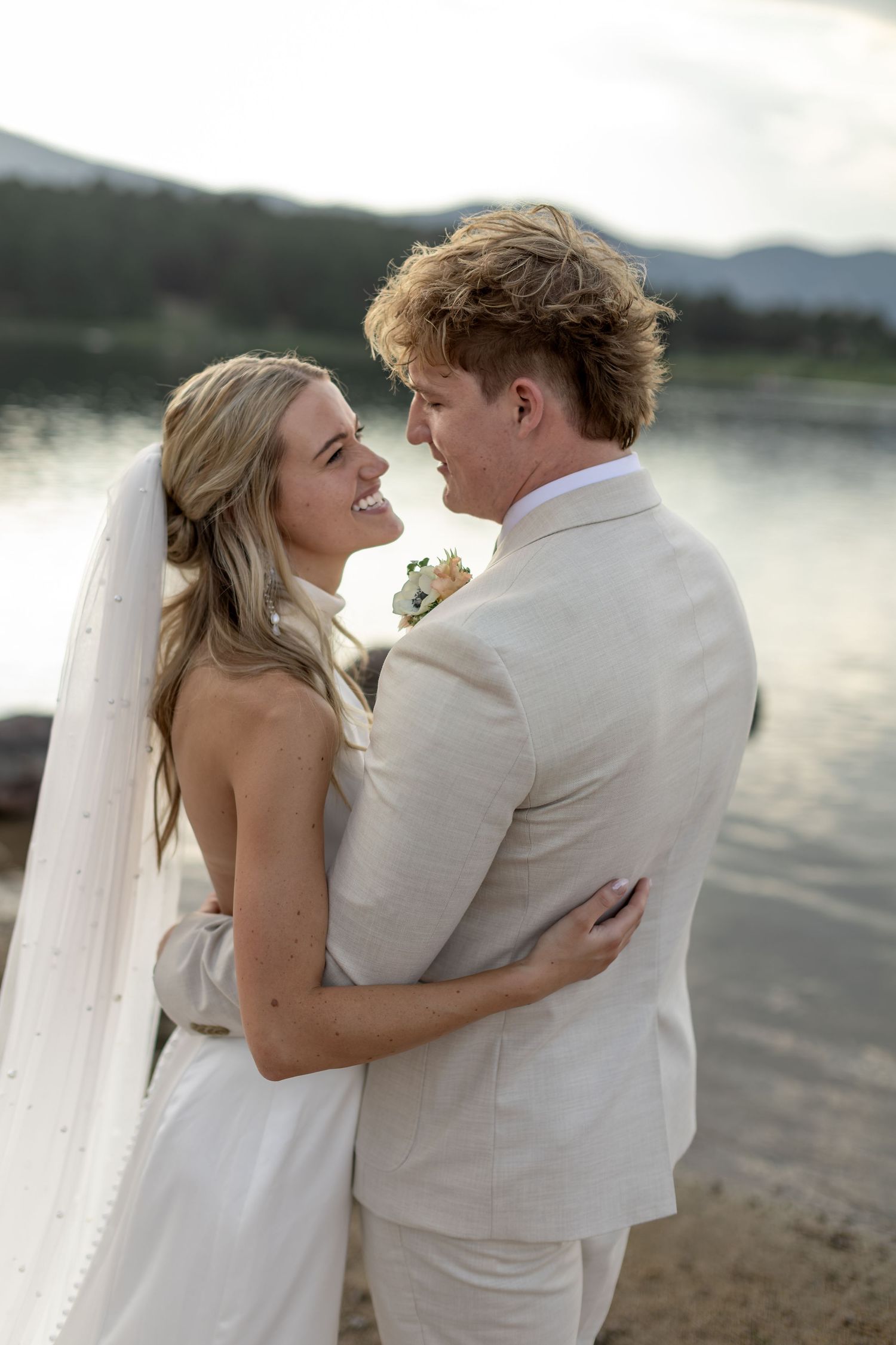 Bride and groom embracing and smiling at each other by a lakeside at sunset in wedding attire.