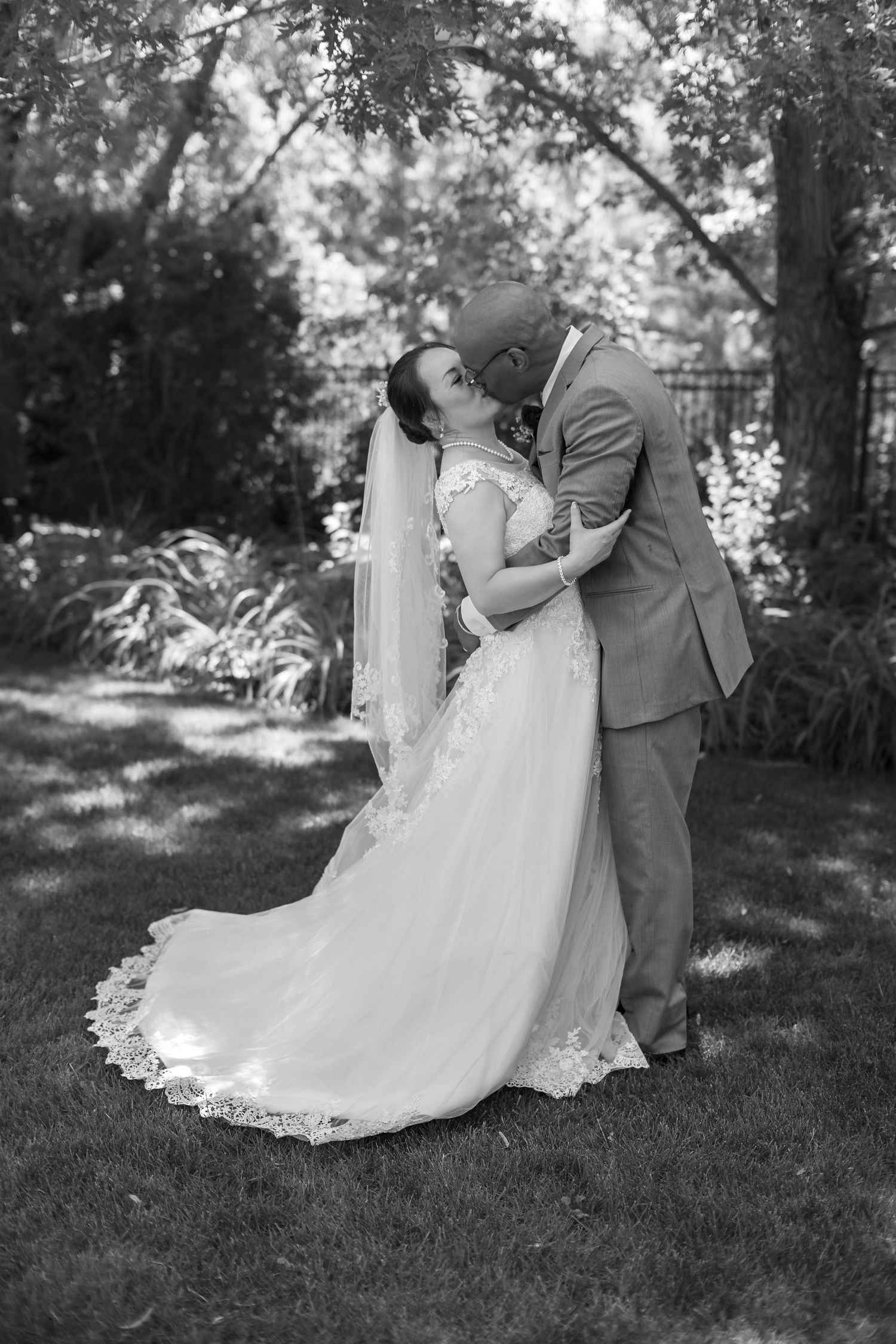 Interracial bride and groom sharing romantic kiss in garden in black and white wedding photo.