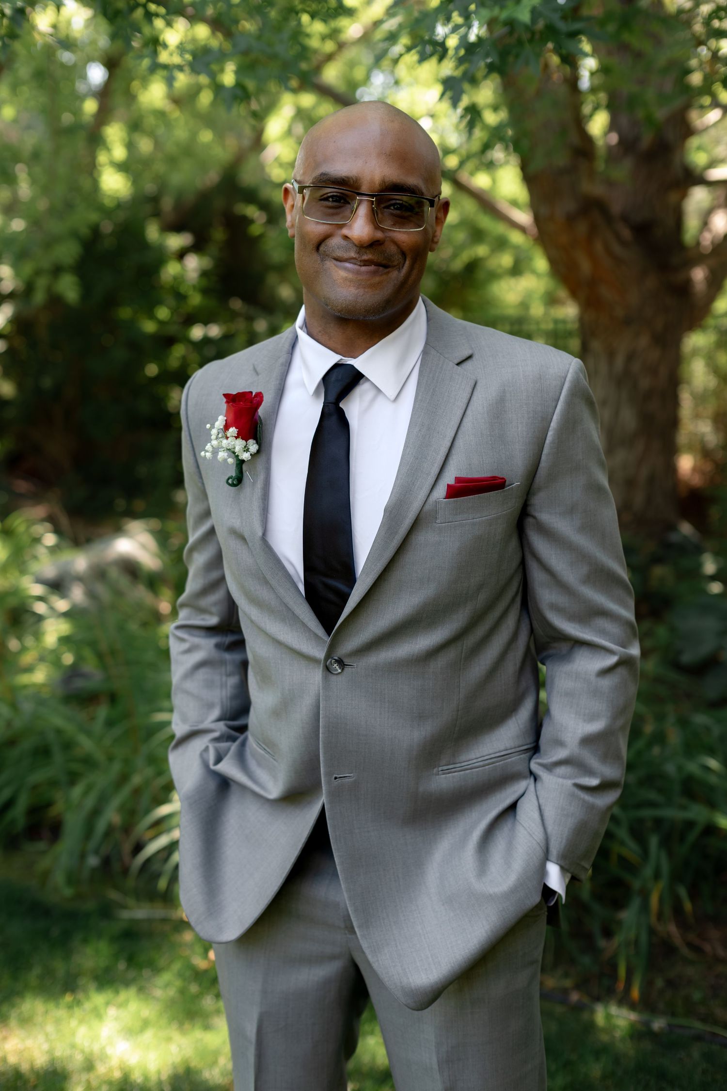 Bald man in gray suit with red boutonniere and pocket square, black tie, smiling outdoors at a wedding.