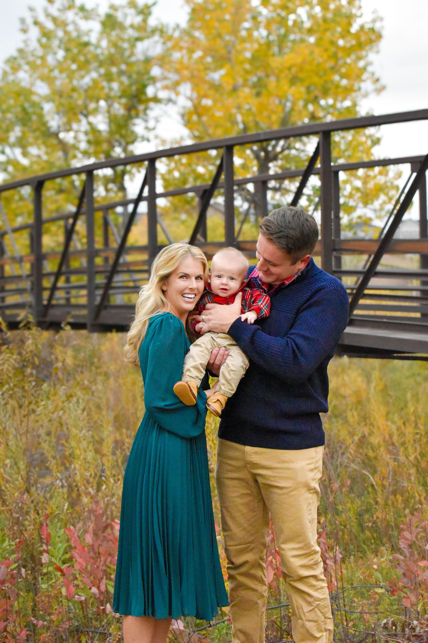 Happy family with baby posing outdoors near a bridge surrounded by colorful autumn foliage.