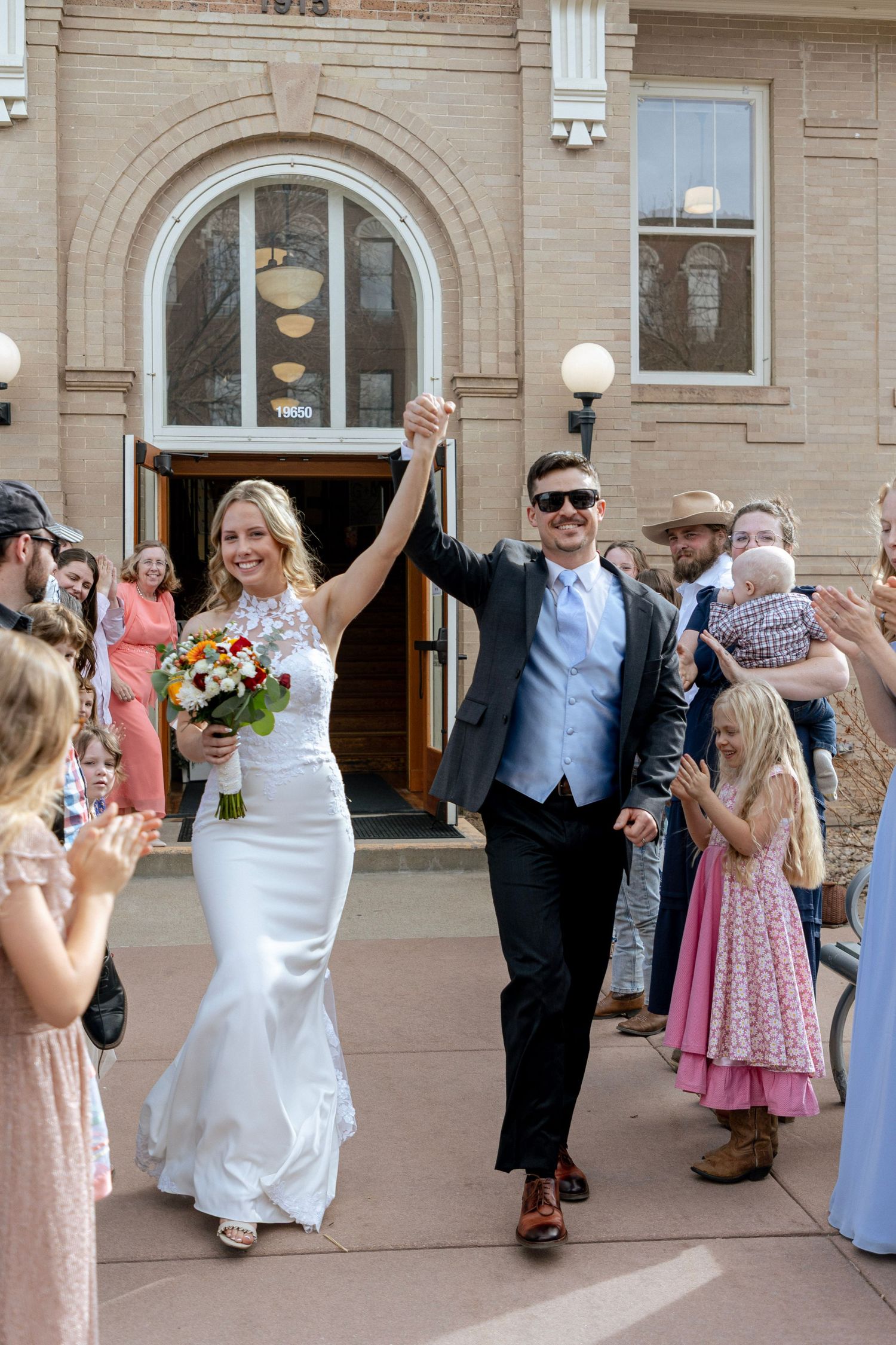 Joyful newlyweds raise hands in celebration outside historic brick building after wedding ceremony.