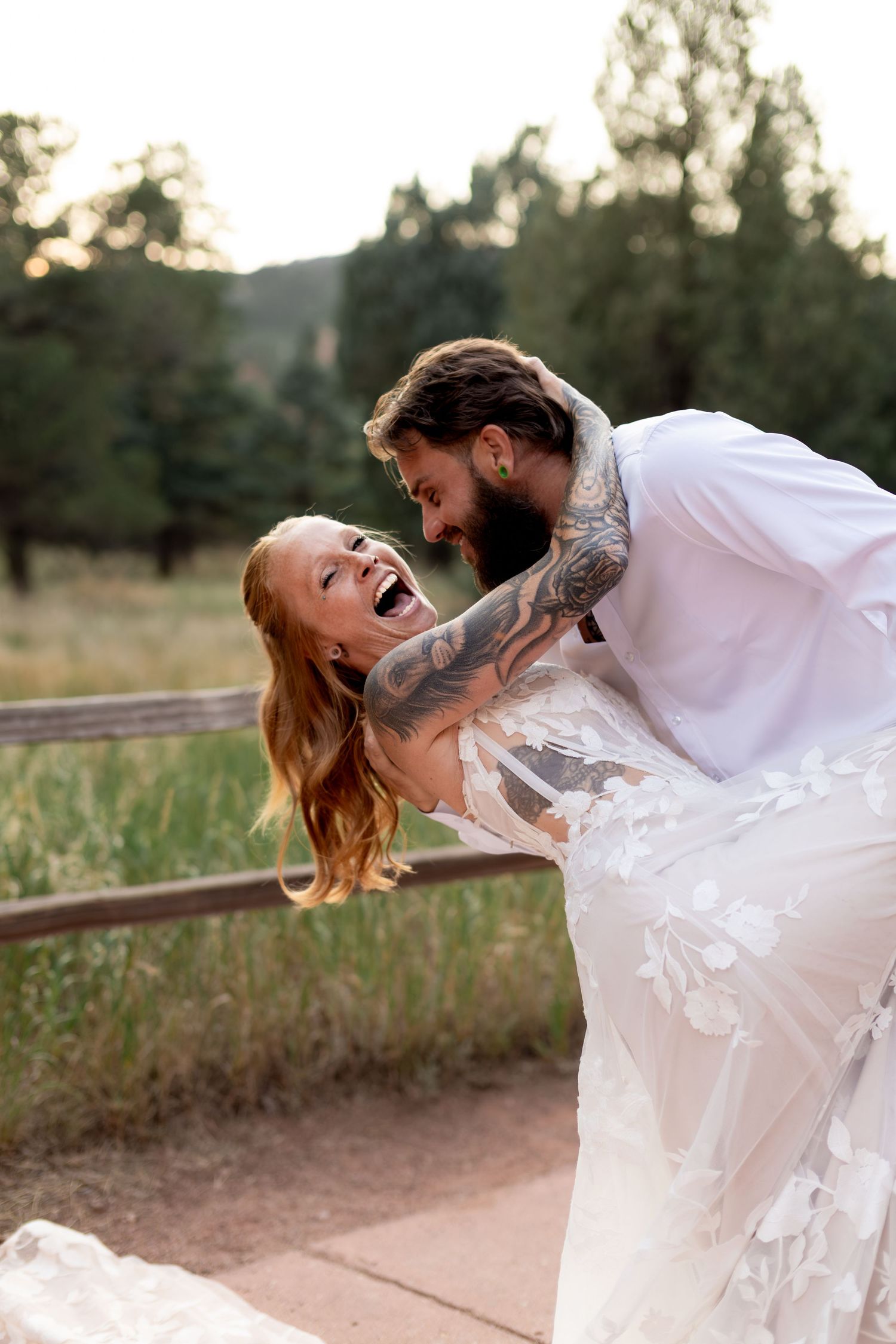 Laughing bride in floral gown being dipped by tattooed groom in white shirt at outdoor wedding.
