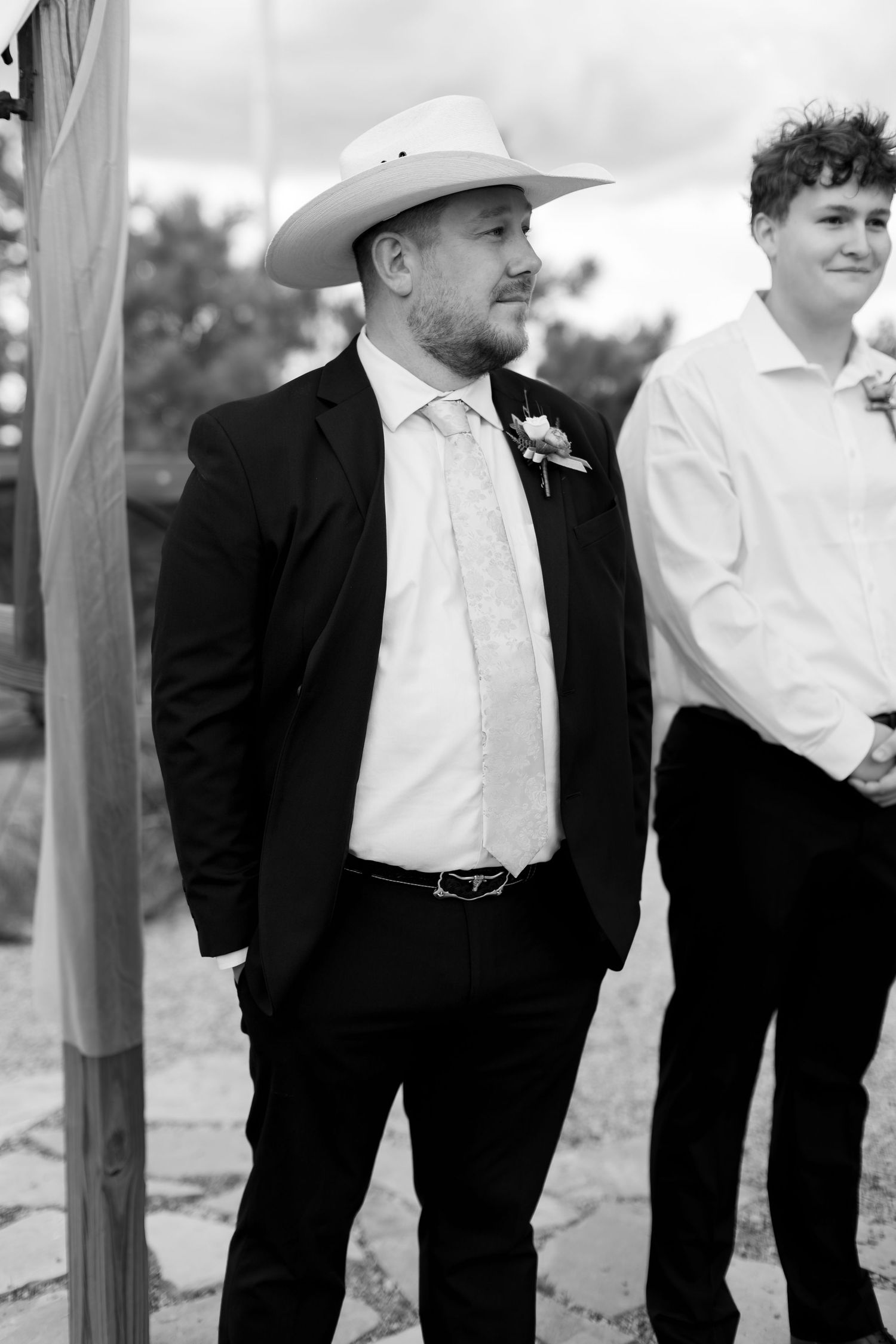 Black and white photo of groom in cowboy hat and black suit with boutonniere at outdoor wedding ceremony.