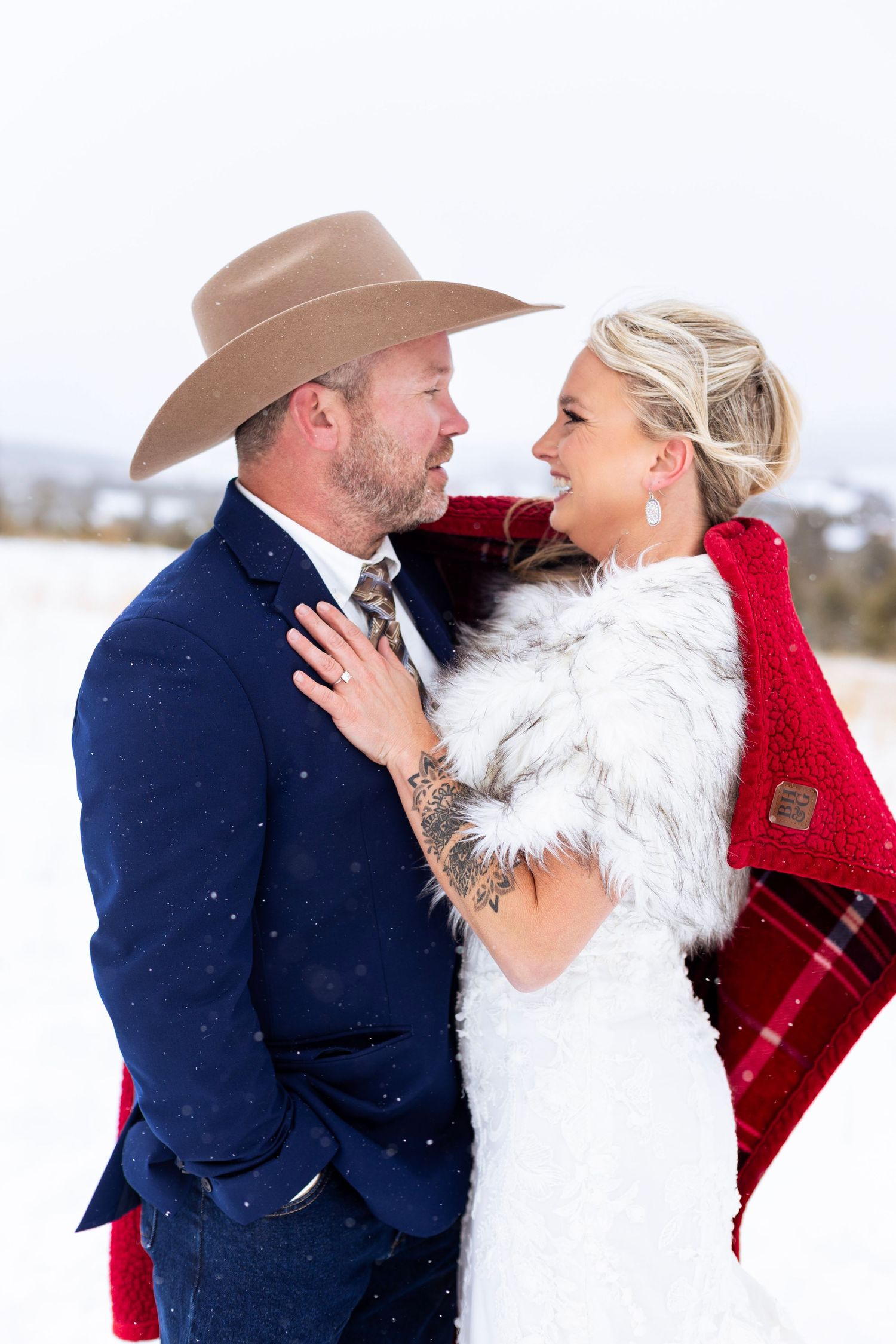 A couple in winter wedding attire share an intimate moment in the snow, with a red plaid blanket and cowboy hat.