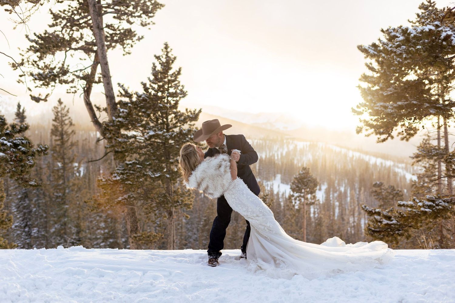 A couple shares a romantic dip pose in the snowy mountains during sunset with evergreen trees in the background.