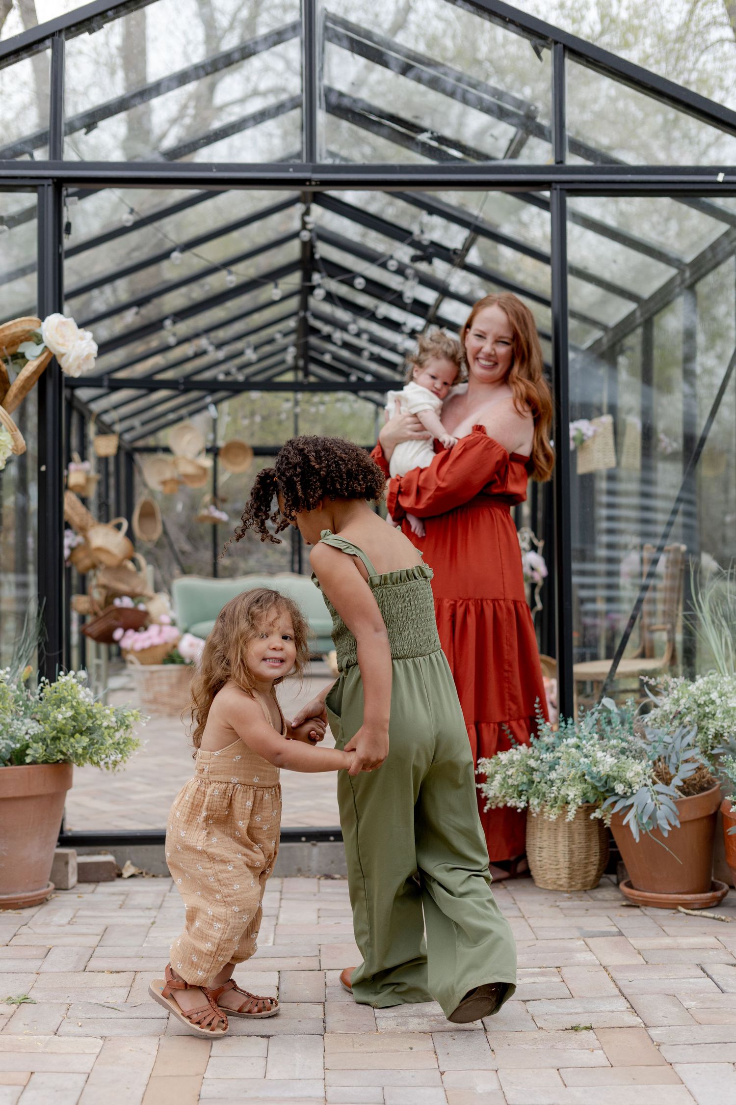 A smiling redhead mother in a red dress holds a baby while two young children play together outside a glass greenhouse.