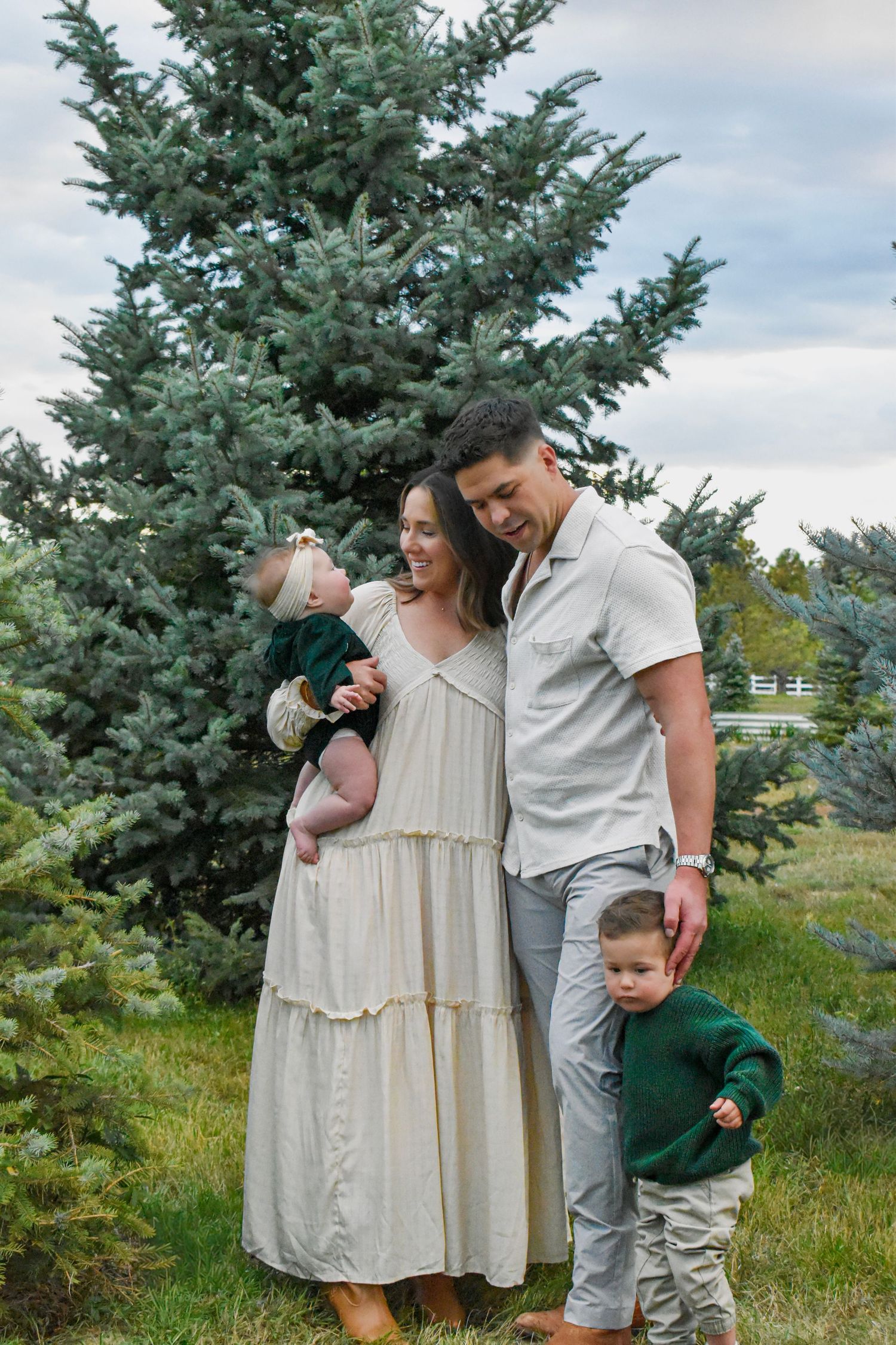 A family in coordinated neutral outfits poses together near a large evergreen tree.