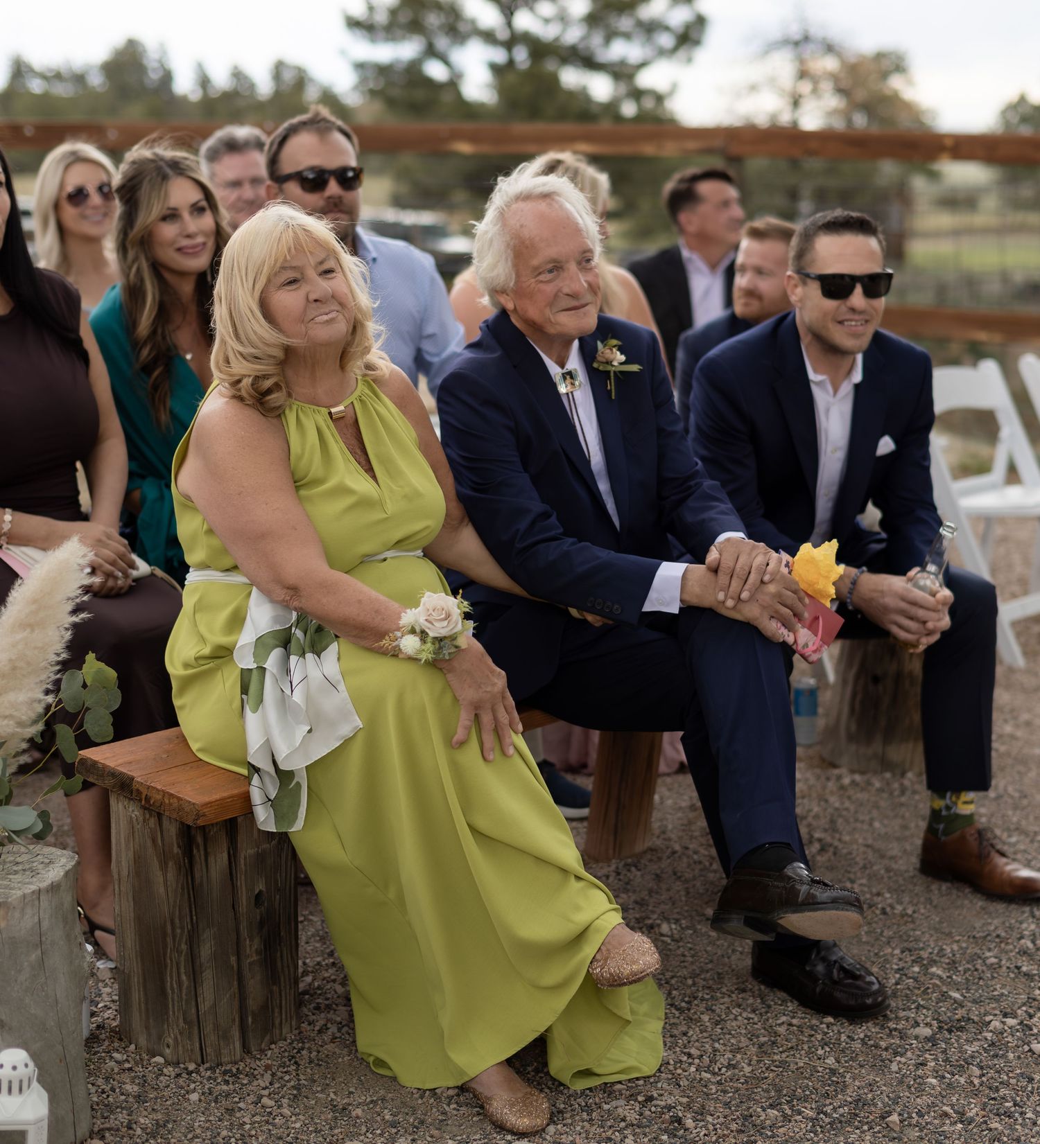 Wedding guests seated outdoors at a rustic ceremony, including a woman in a green dress.