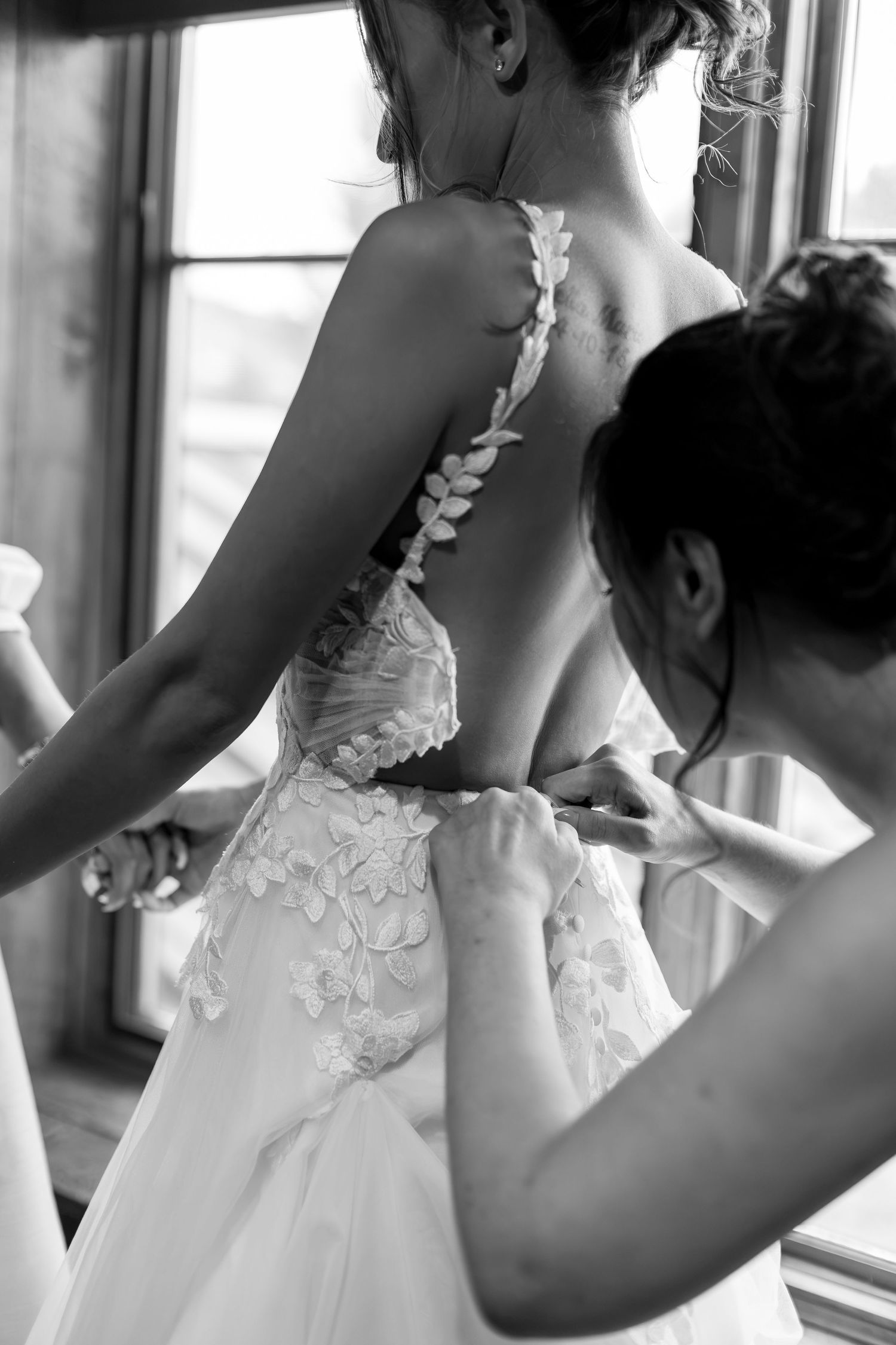 Black and white photo of a bride having her open-back floral wedding gown buttoned up by a bridesmaid.