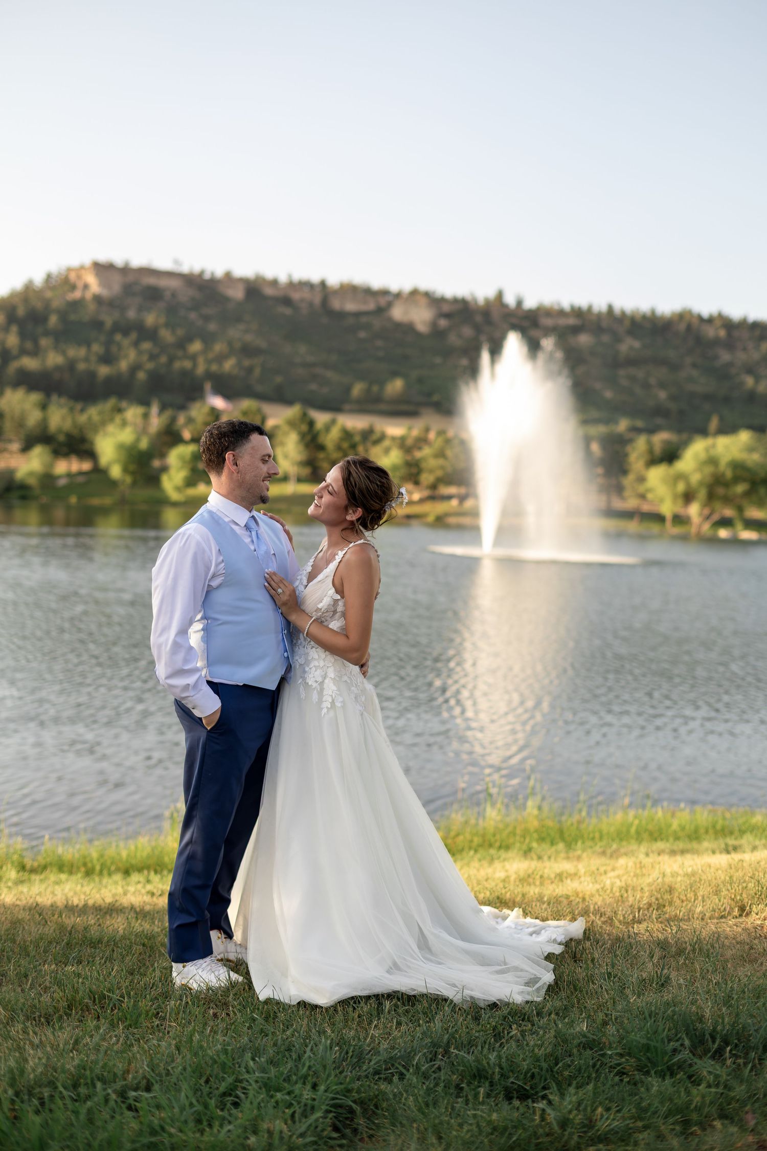 Bride and groom embracing by a lake with a fountain and mountain backdrop at sunset.