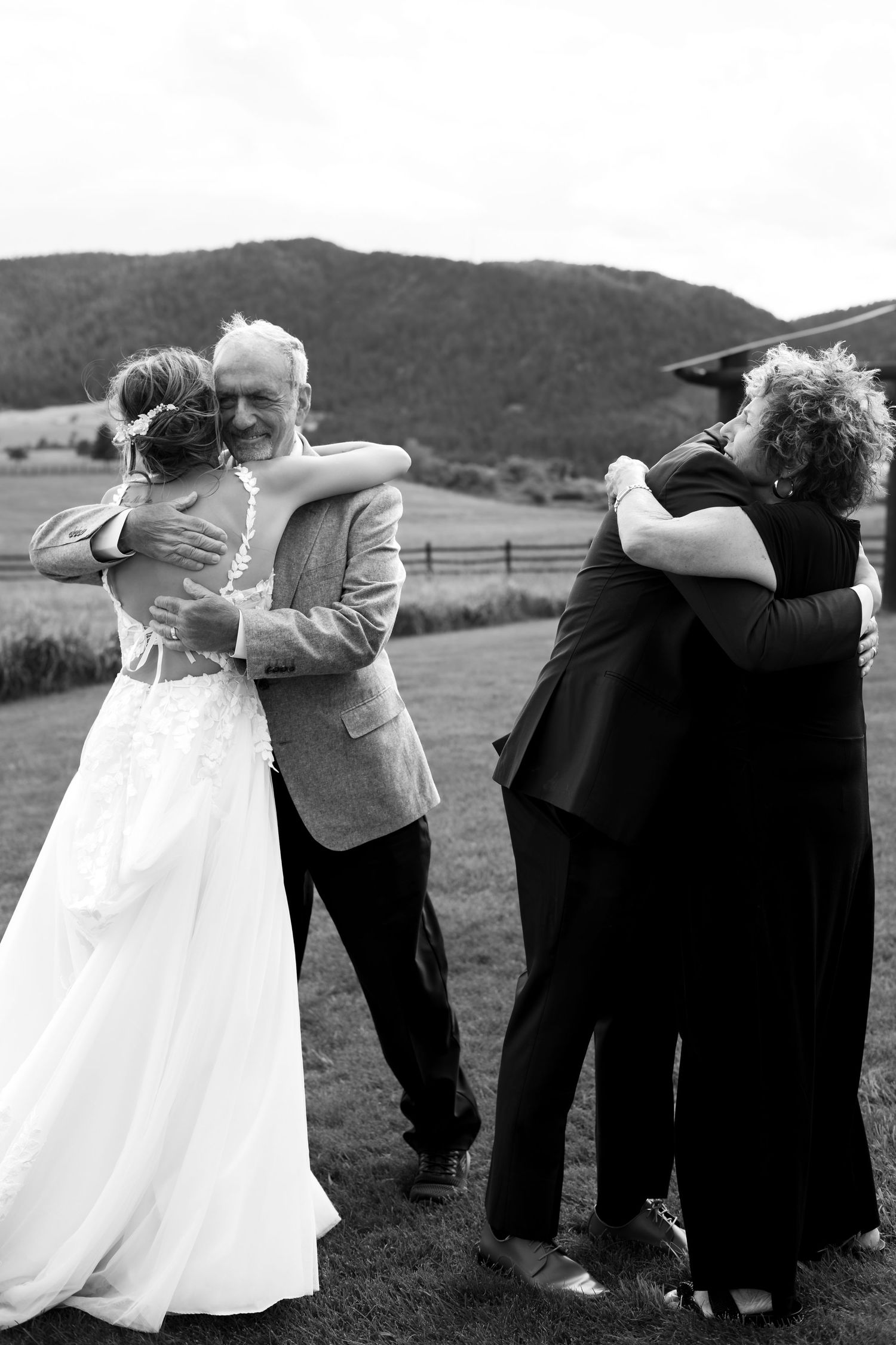 Black and white photo of a bride and groom sharing emotional hugs with family members outdoors at a wedding.