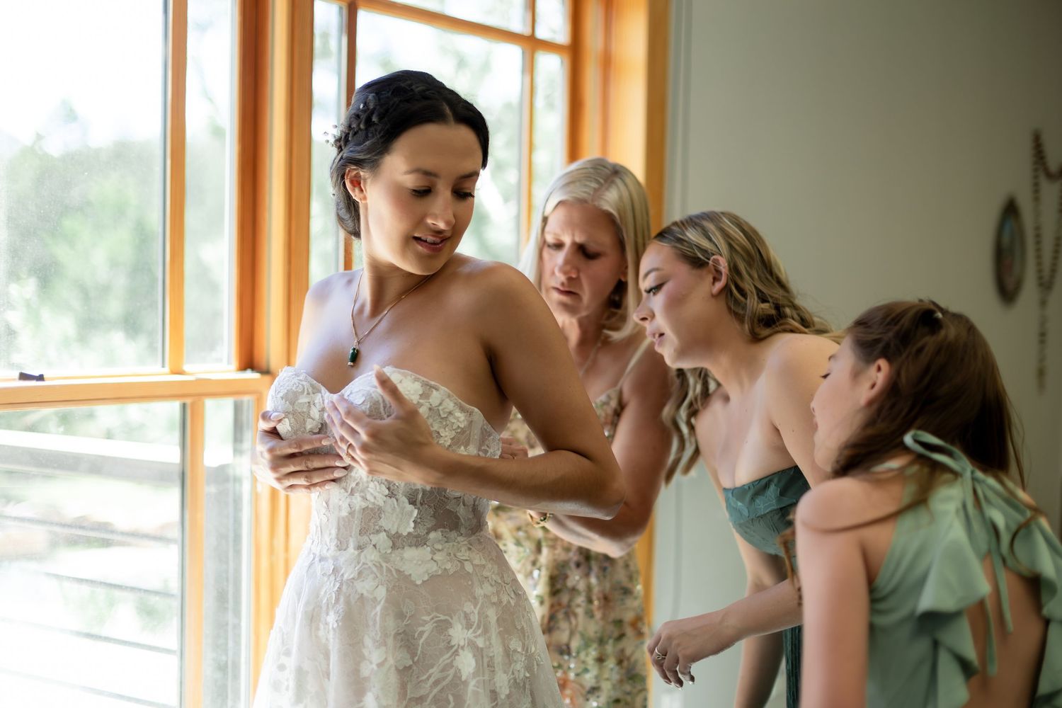 Bride in white lace gown being helped by bridesmaids in sage green dresses near a bright window.