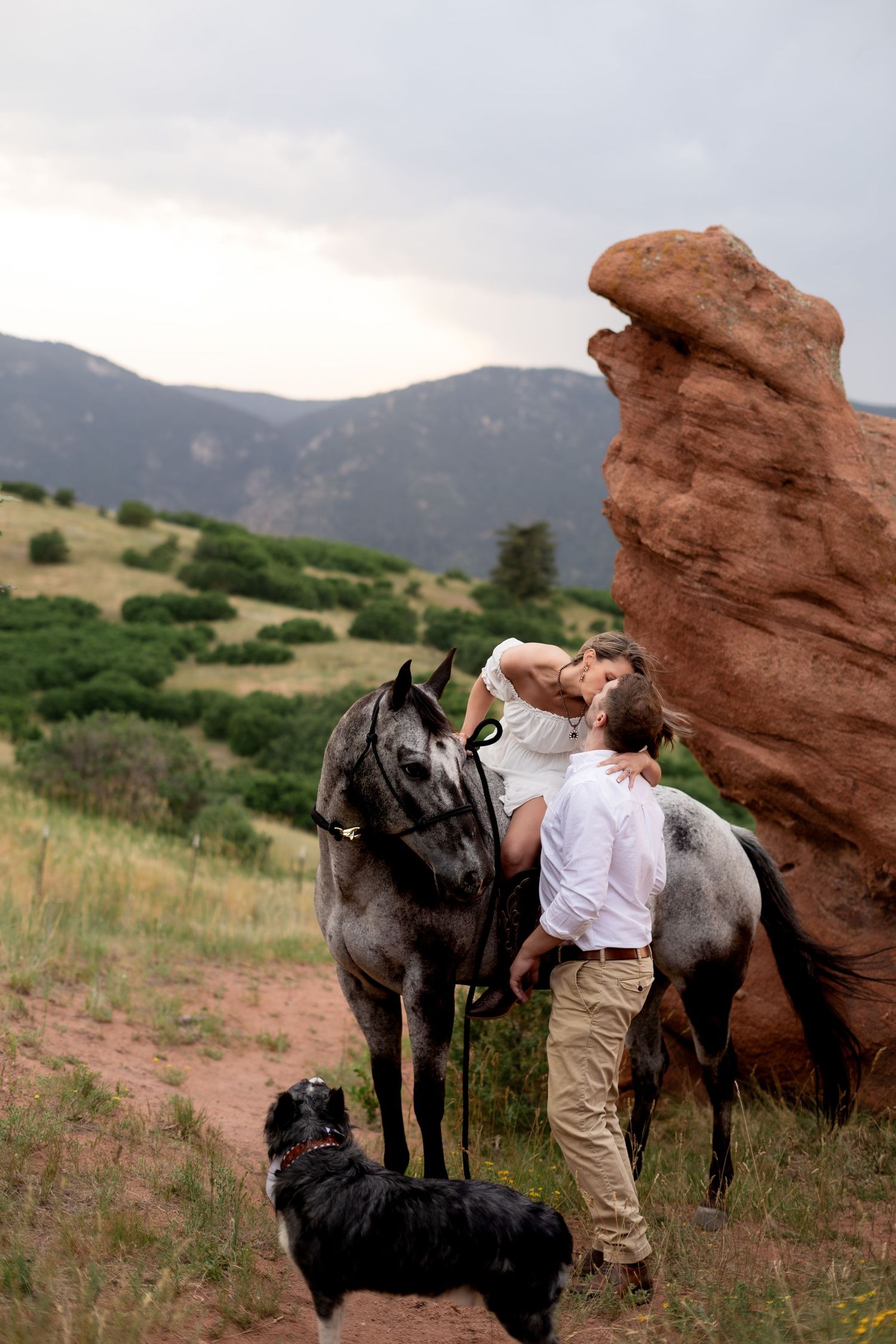 Couple kissing near red rock formations with two gray horses and a black dog in a scenic mountain landscape.