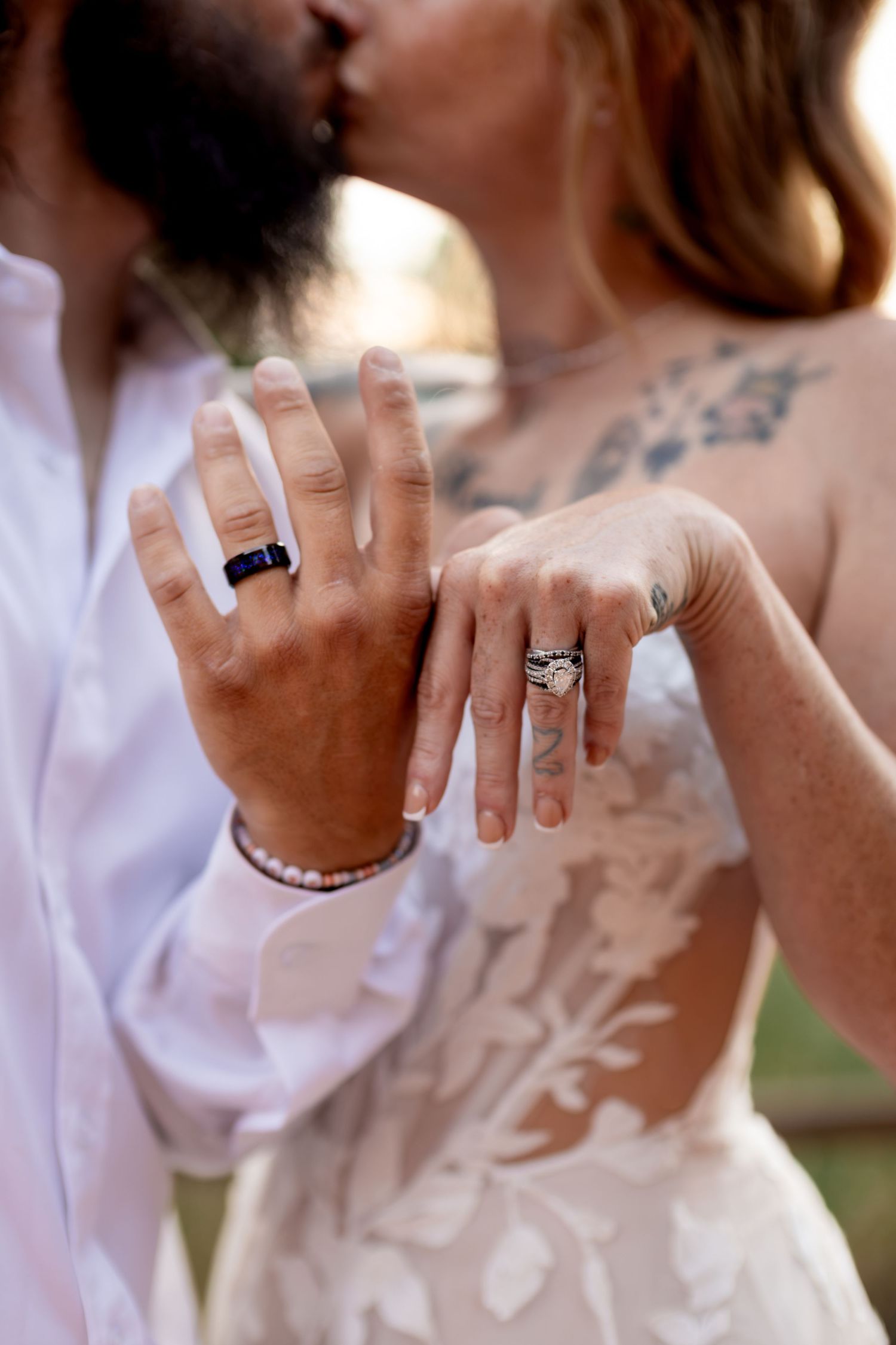 Newlyweds kissing while showing off their wedding rings, bride with floral tattoos in lace gown.