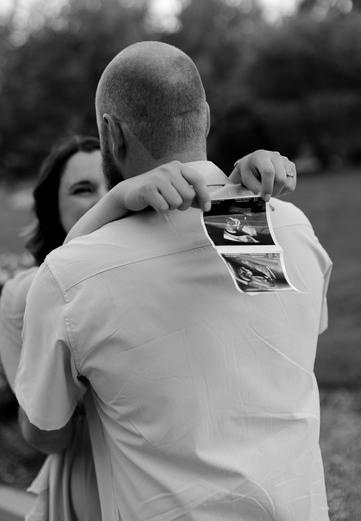 Black and white photo of couple embracing outdoors, woman holding ultrasound photos behind mans back, pregnancy announcement.