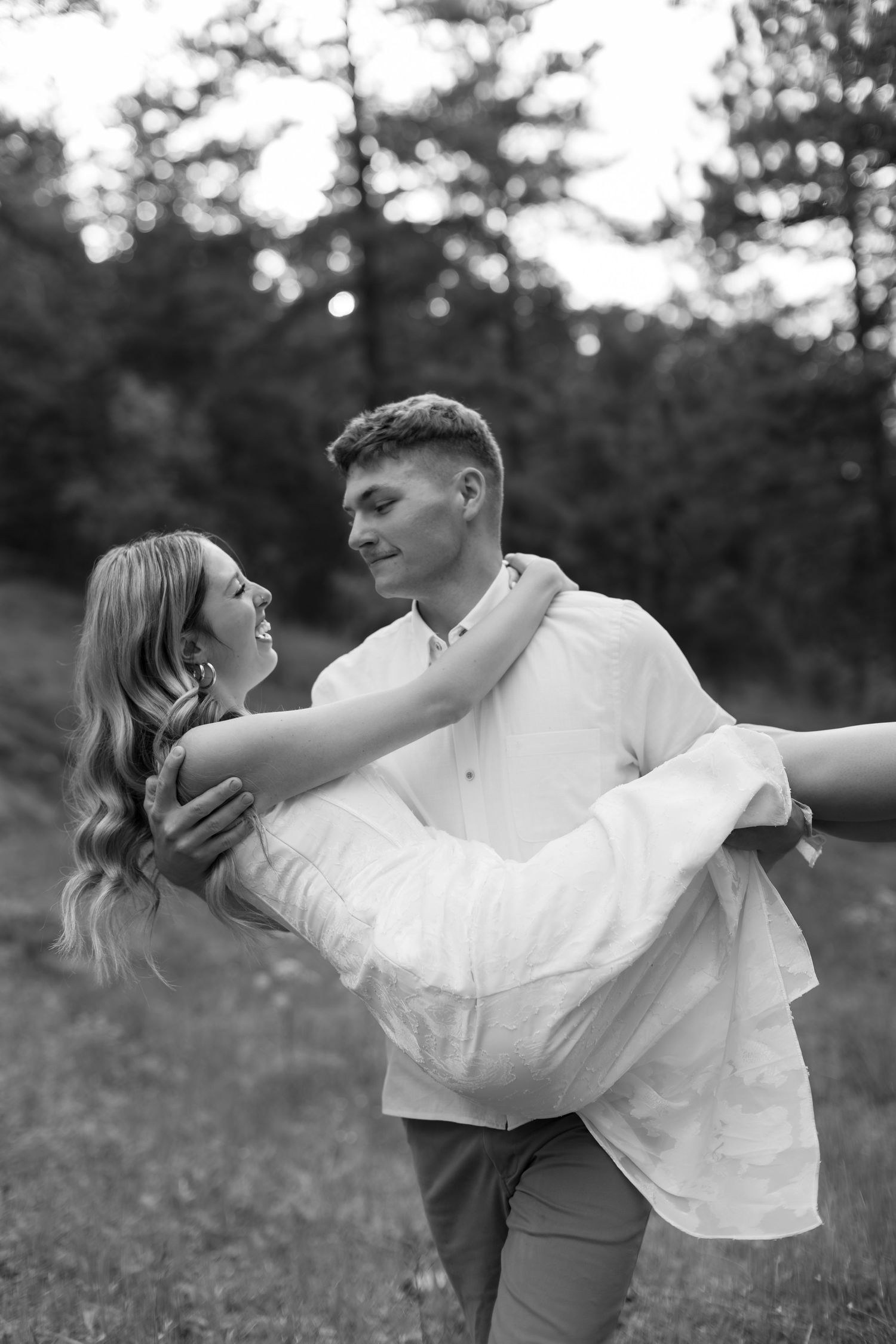 Black and white photo of a man carrying a woman in his arms while gazing at each other in a lush outdoor setting.