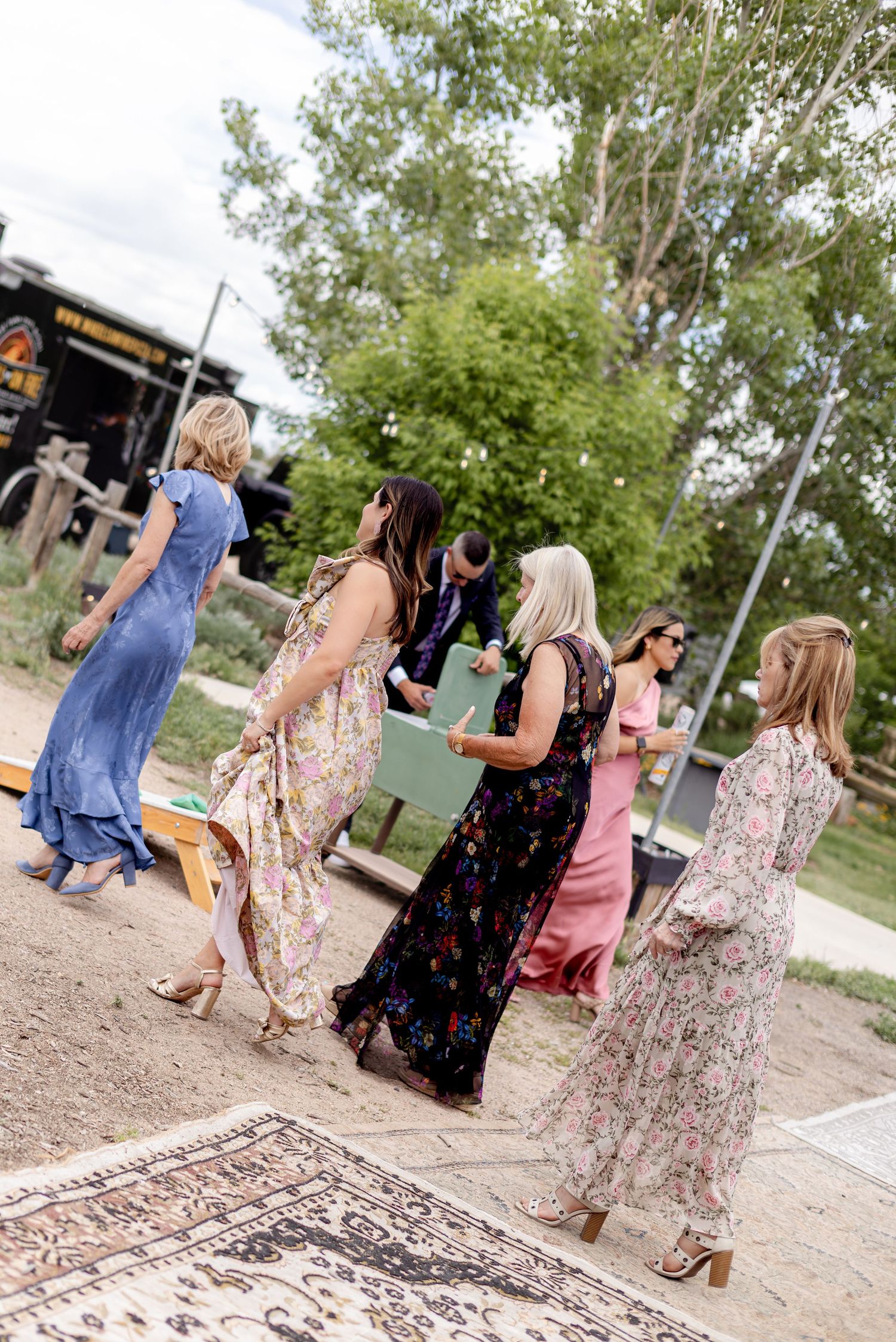 Wedding guests in colorful floral and blue maxi dresses mingling outdoors near rustic venue with trees.