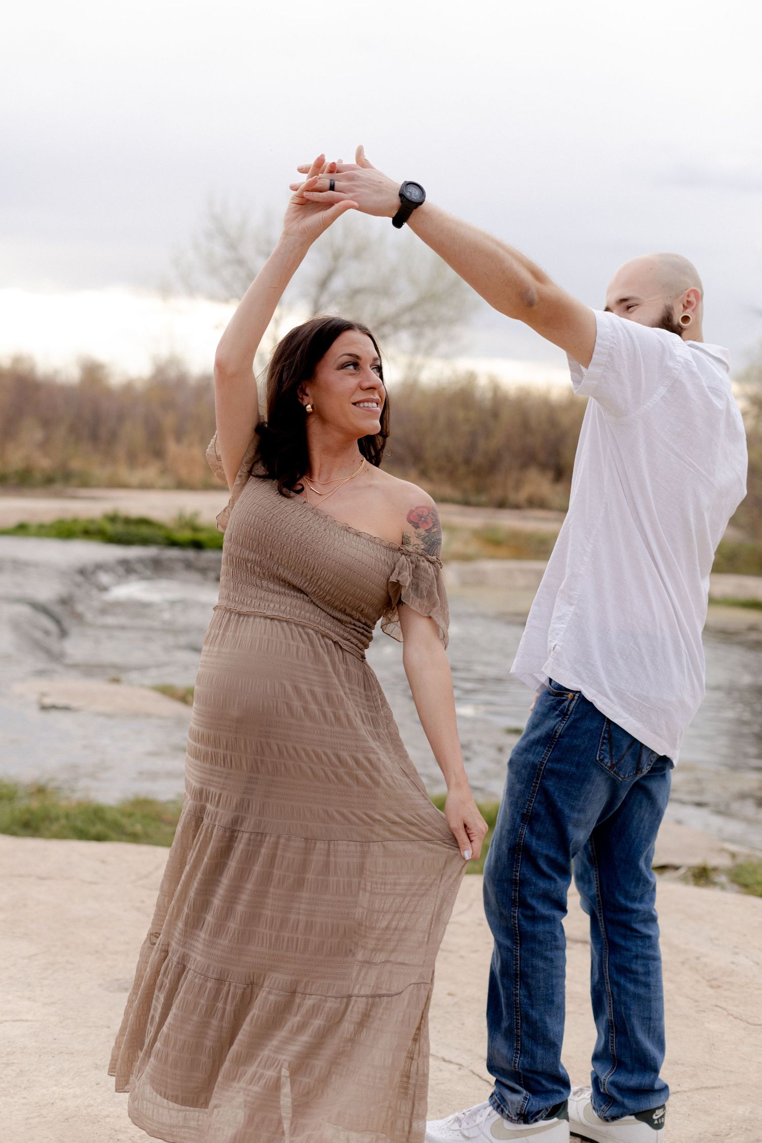 A couple dancing outdoors near a river, woman in tan dress smiling as man in white shirt twirls her around.