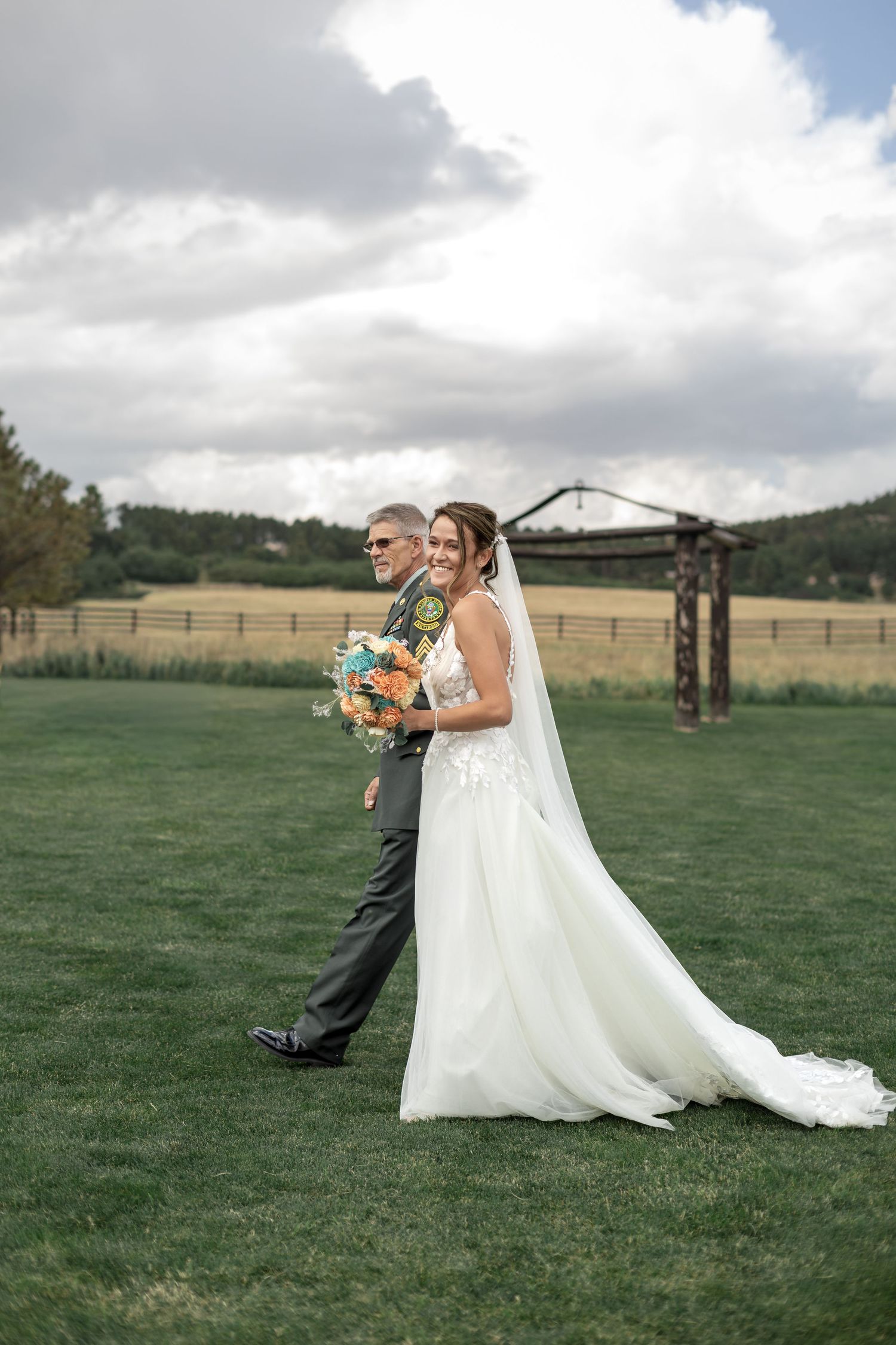 Bride in lace gown with colorful bouquet walks with father across green field on wedding day.