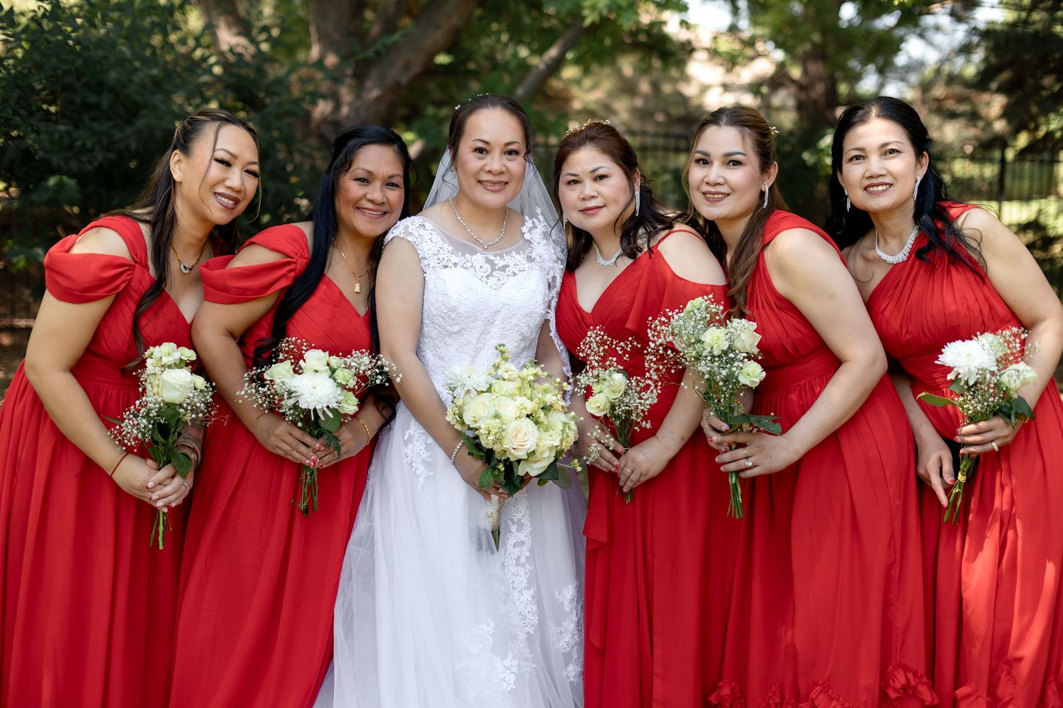 Bride in white gown with five bridesmaids in red dresses holding bouquets outdoors.
