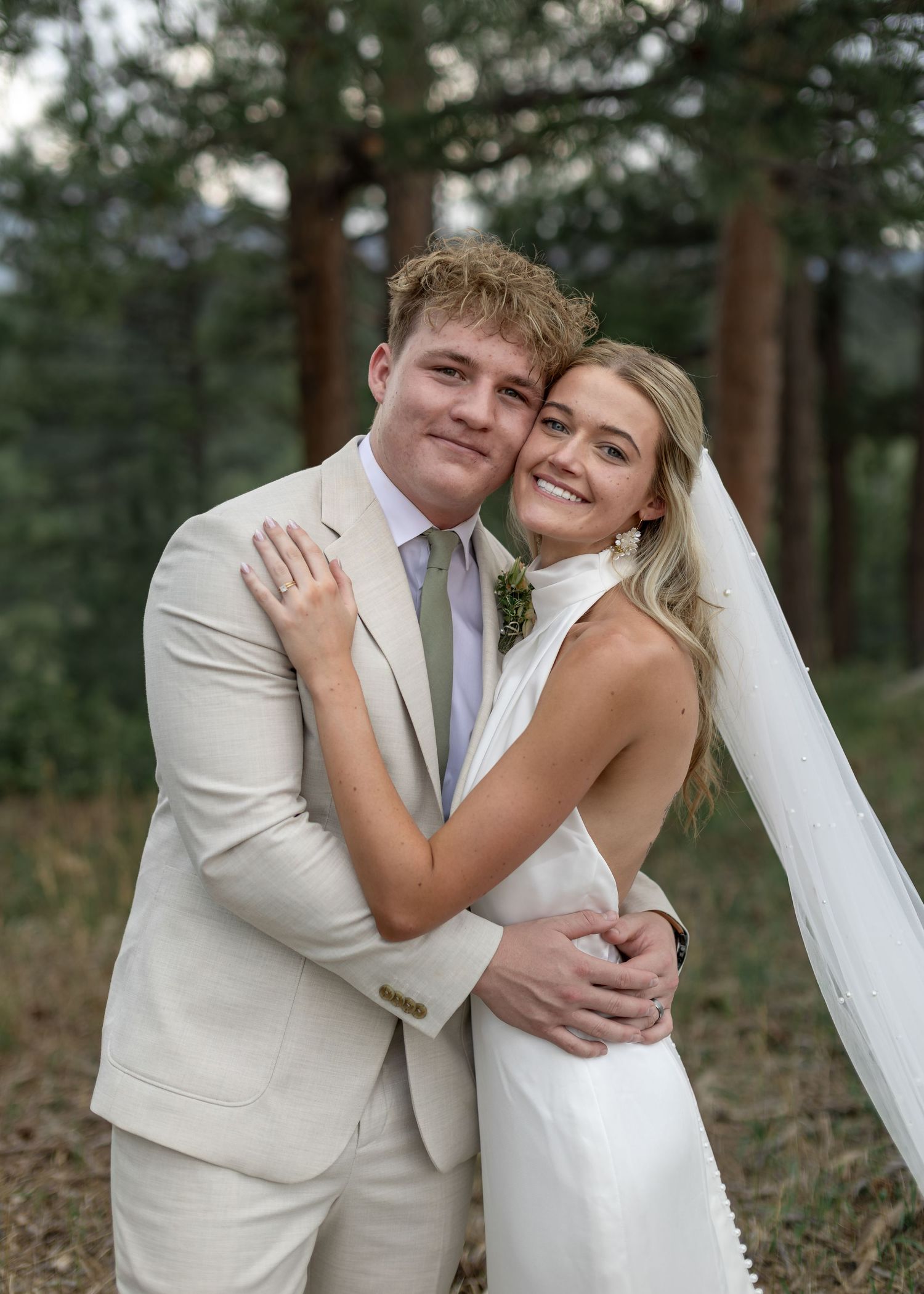 Smiling bride and groom hugging outdoors among pine trees, bride in white halter dress with veil.