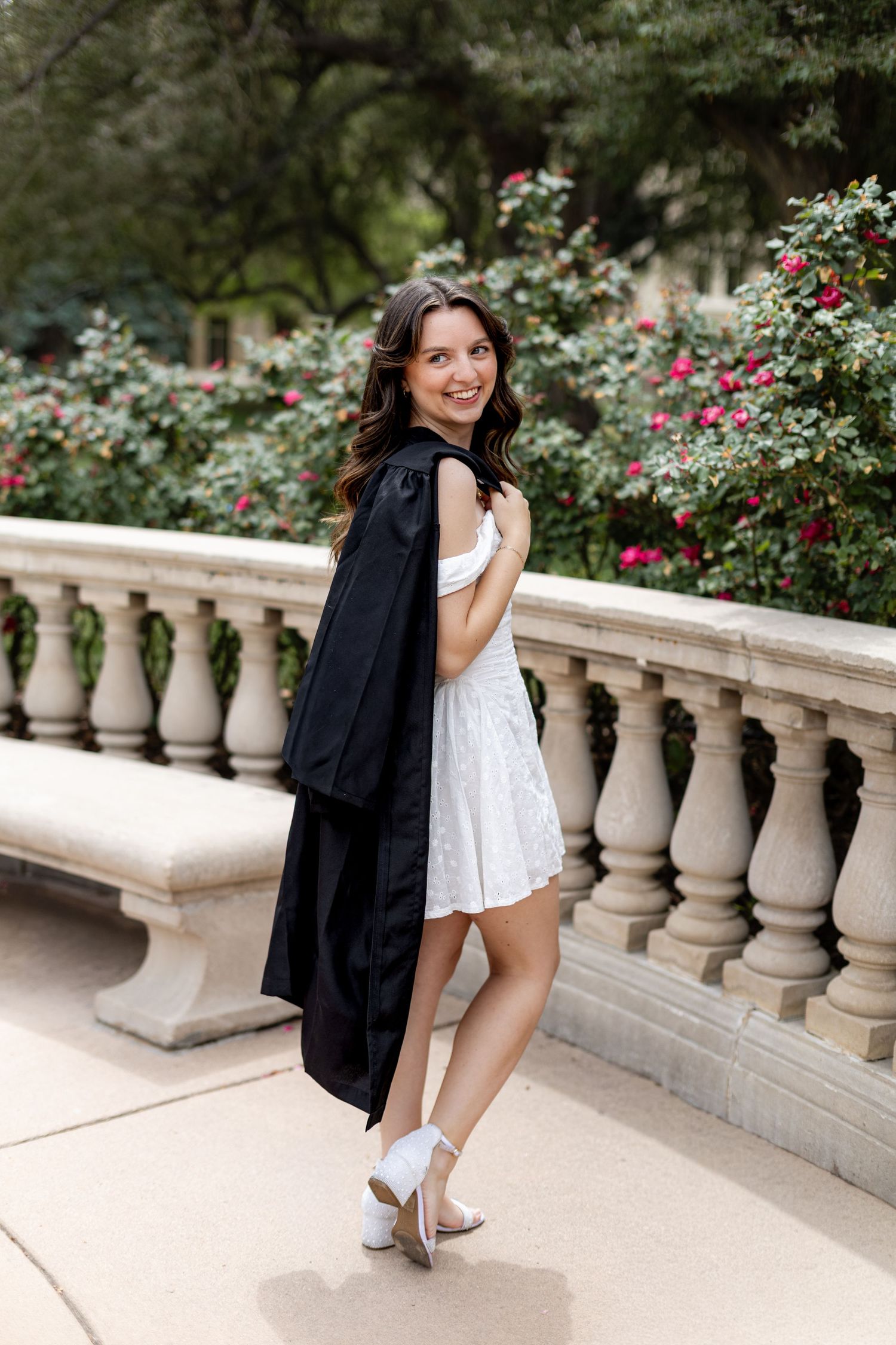 Smiling young woman in white dress holding black graduation gown, posing by stone balustrade with flowering bushes.