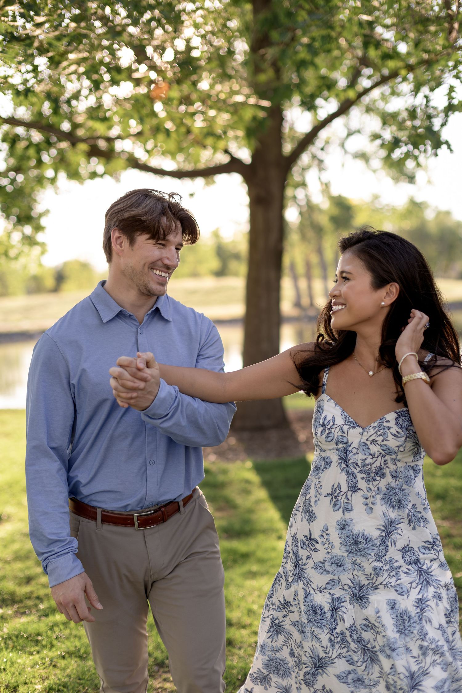 A smiling couple dances outdoors in a park, man in blue shirt and woman in floral dress, holding hands under green trees.