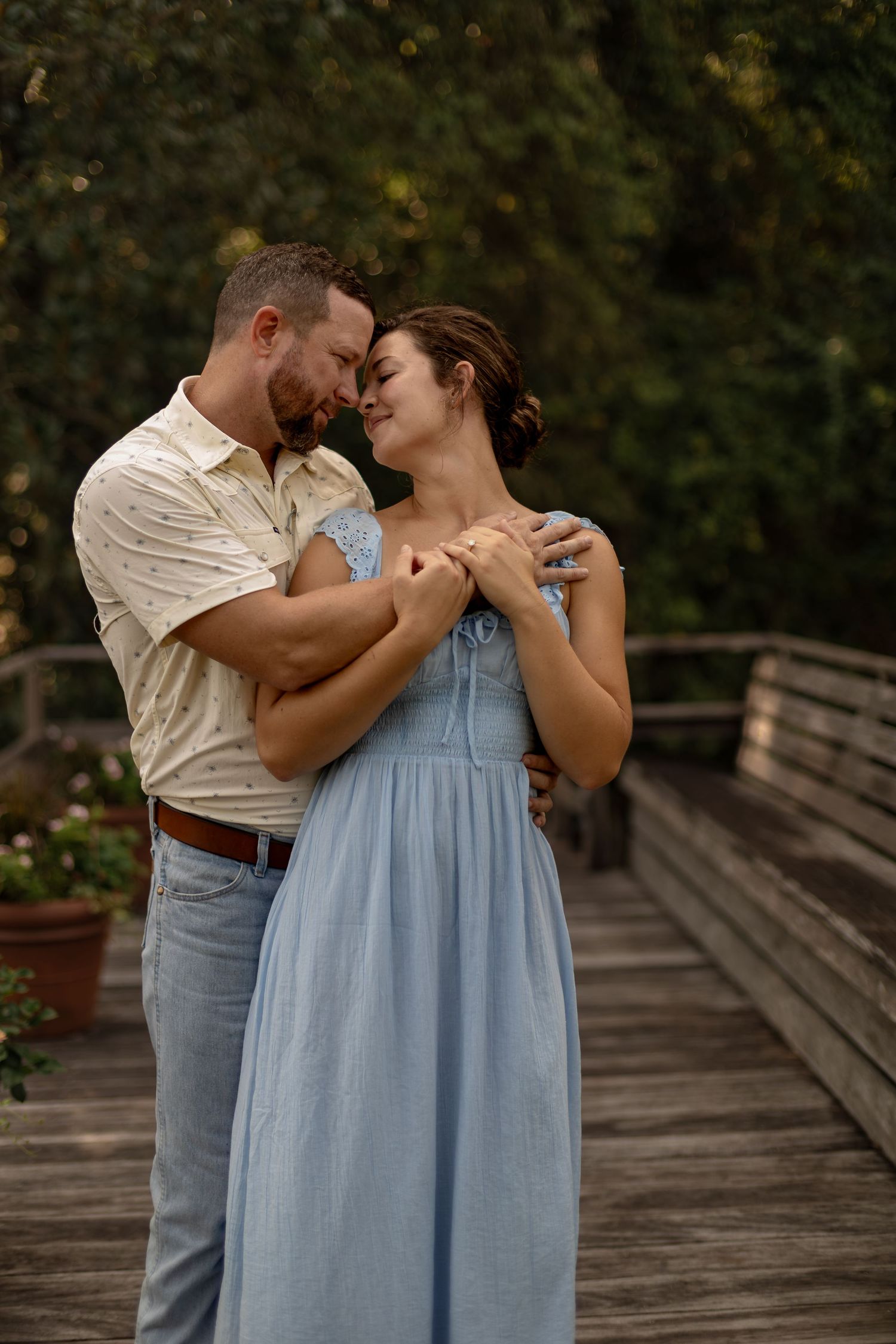 Engaged couple embraces tenderly on a wooden bridge surrounded by lush green trees.