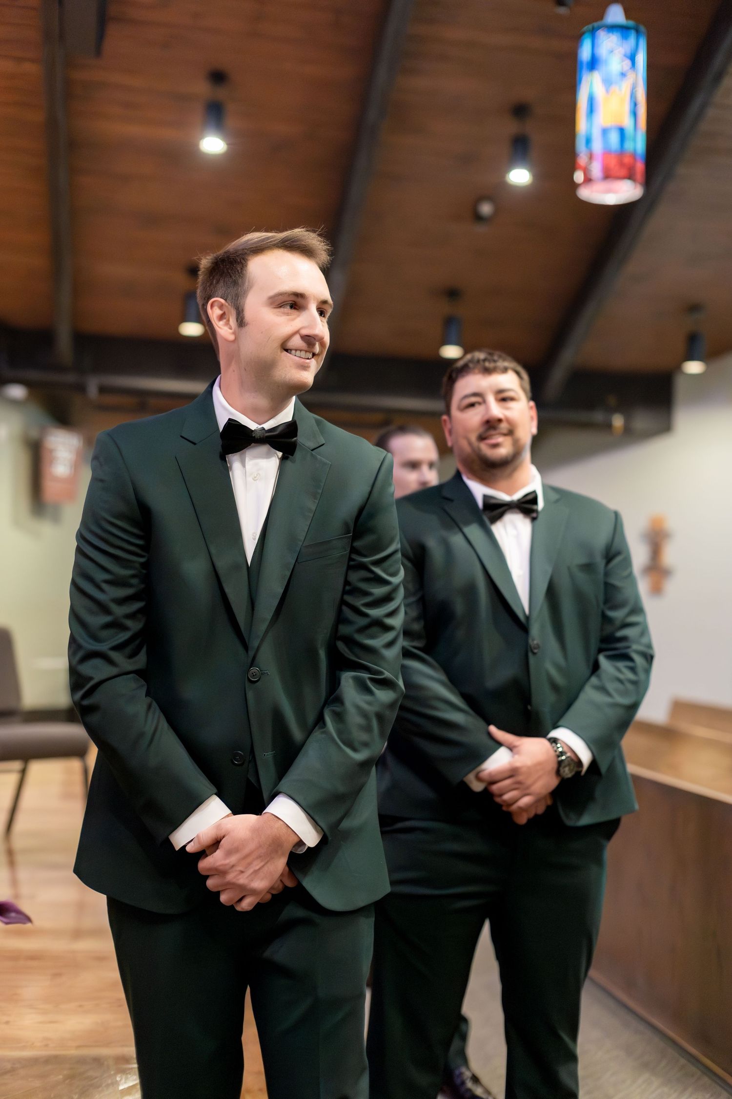 Two groomsmen in dark green tuxedos with black bow ties smile during a wedding ceremony in a wood-paneled church.