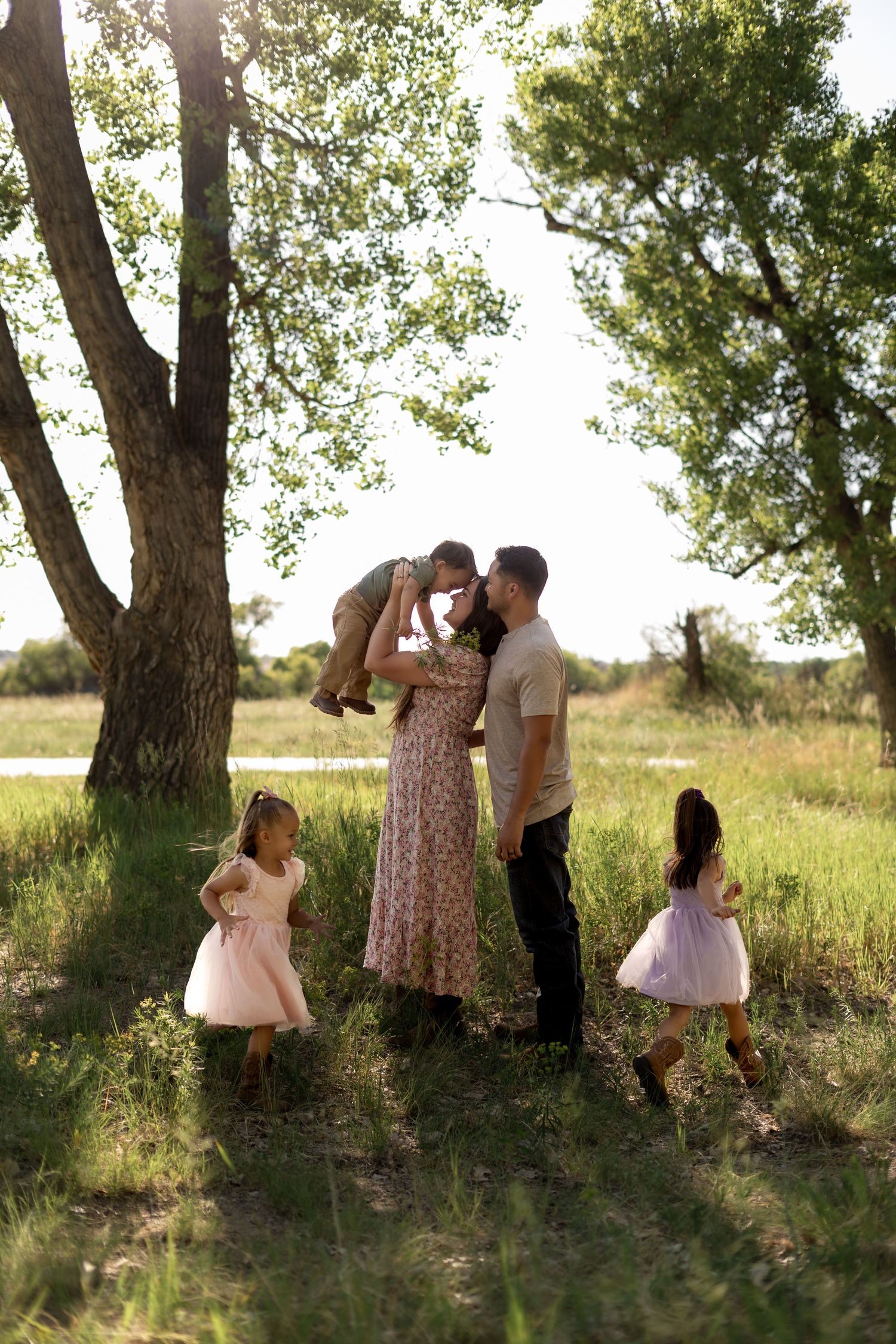 A happy family with two girls in pink dresses enjoys a sunny outdoor moment as parents lift their young son in a green park.