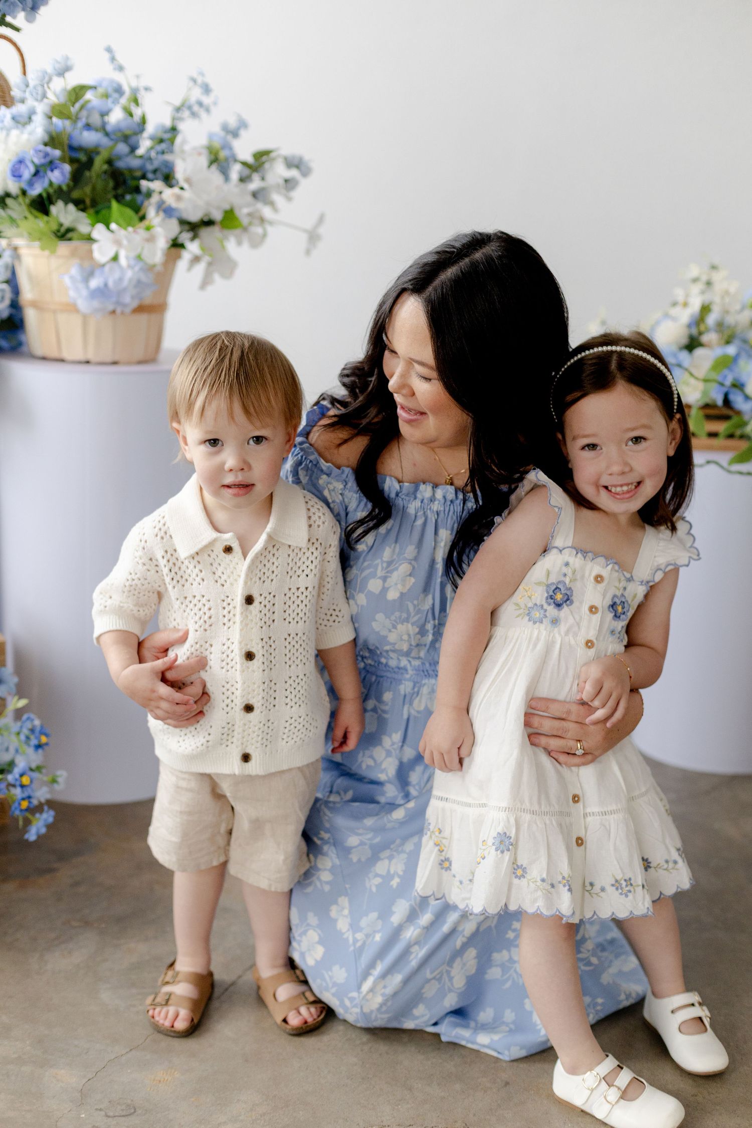 A mother in a blue floral dress poses with her young son and daughter surrounded by blue hydrangeas in a studio.
