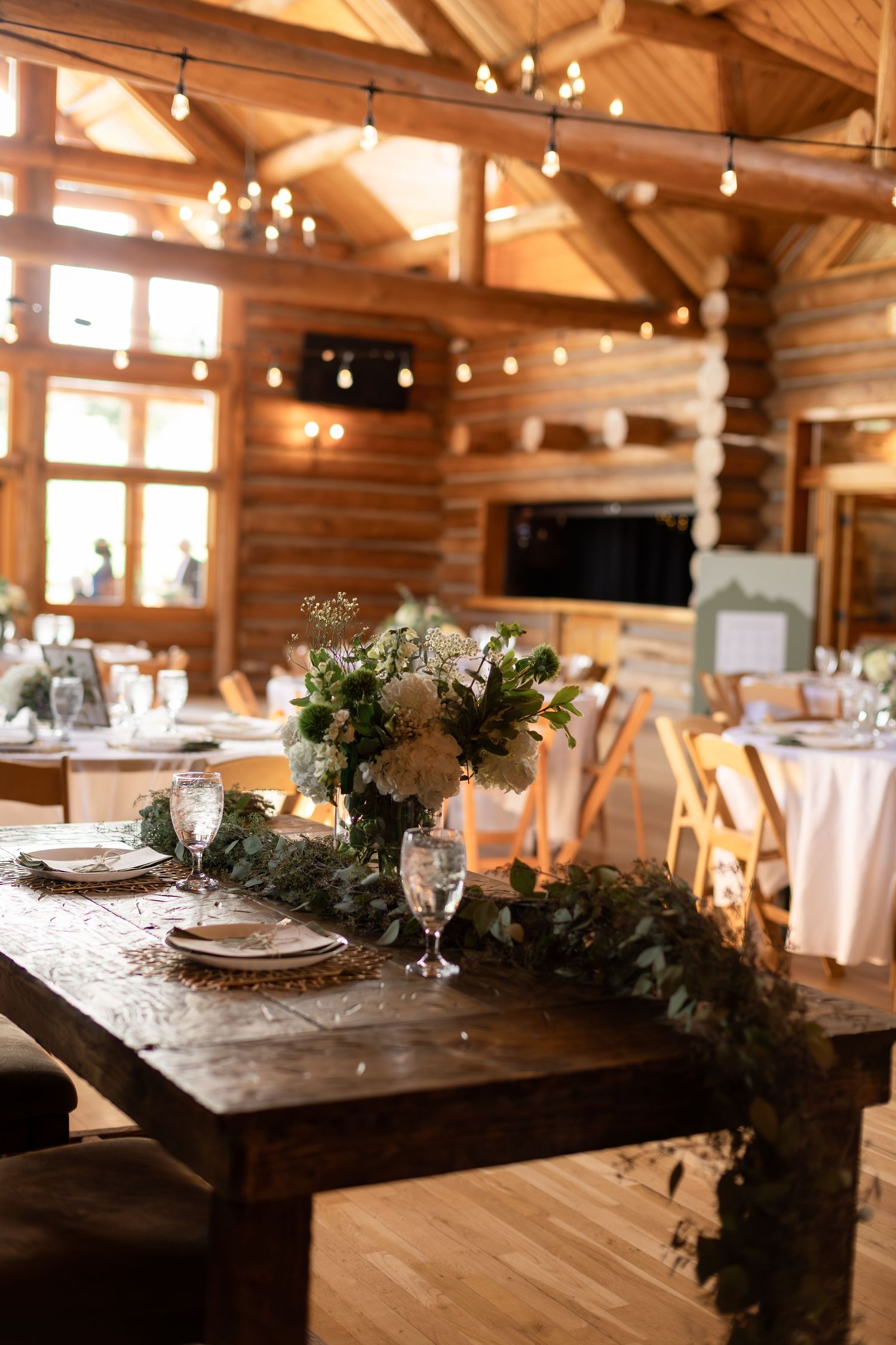 Rustic wedding reception in a log cabin venue with floral centerpieces, greenery table runner, string lights, and elegant place settings.