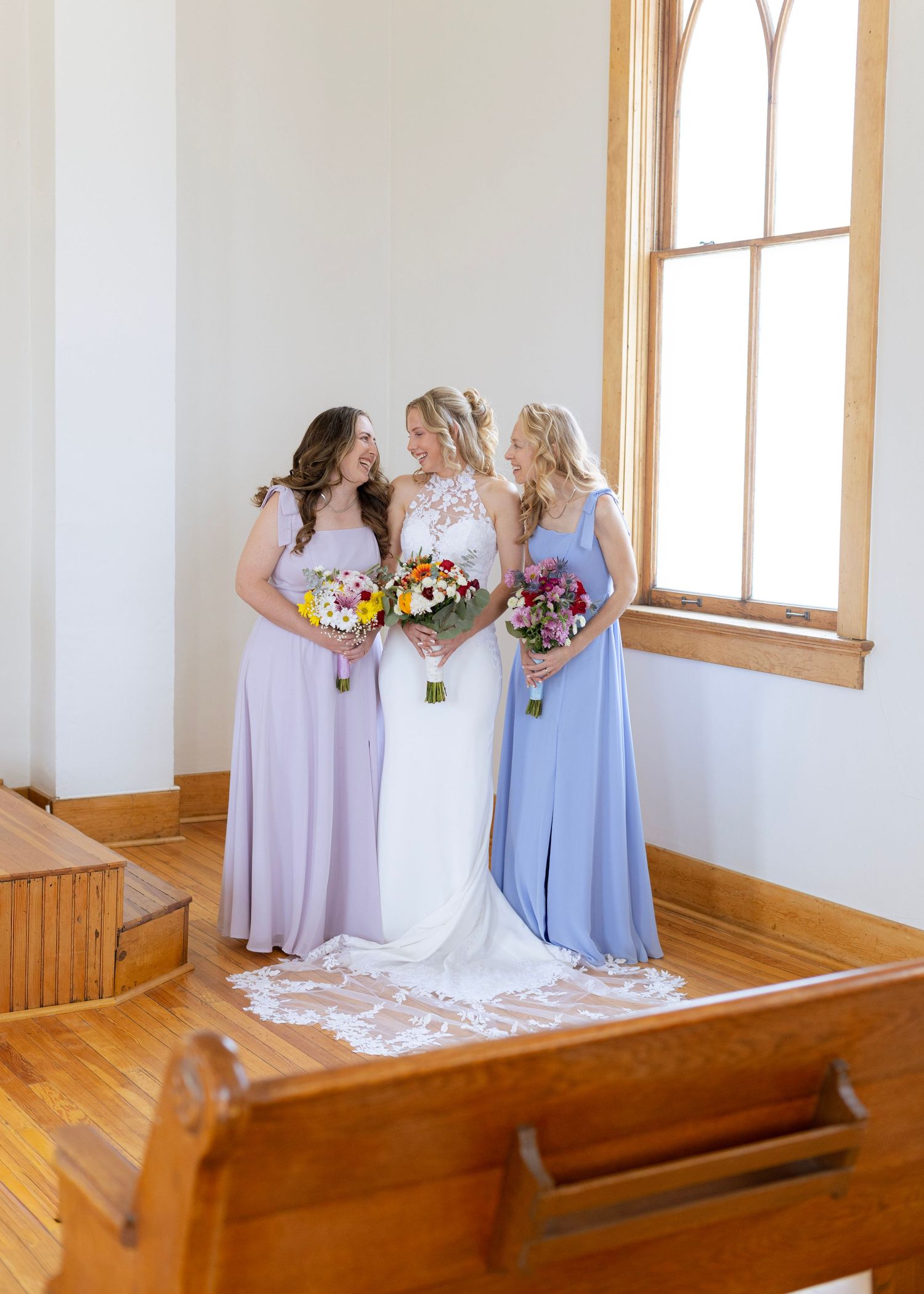 Bride in white lace gown stands with two bridesmaids in lavender dresses holding colorful bouquets inside a bright church.
