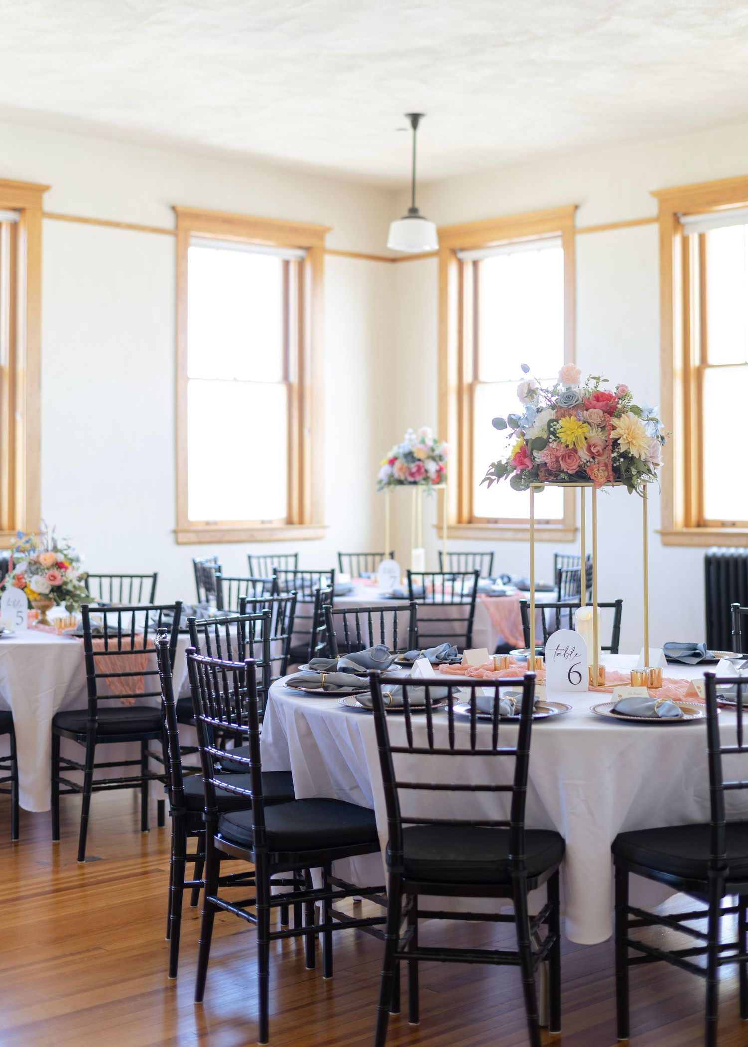 Elegant wedding reception venue with round tables, white linens, black chiavari chairs, and tall floral centerpieces by bright windows.