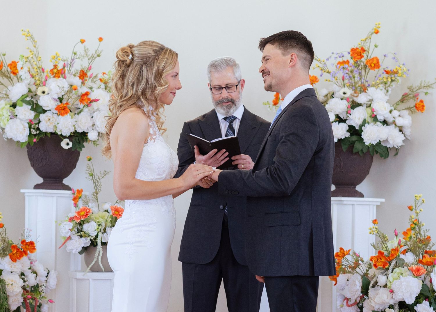 Bride and groom exchange rings at wedding altar with orange and white floral arrangements and officiant behind them.