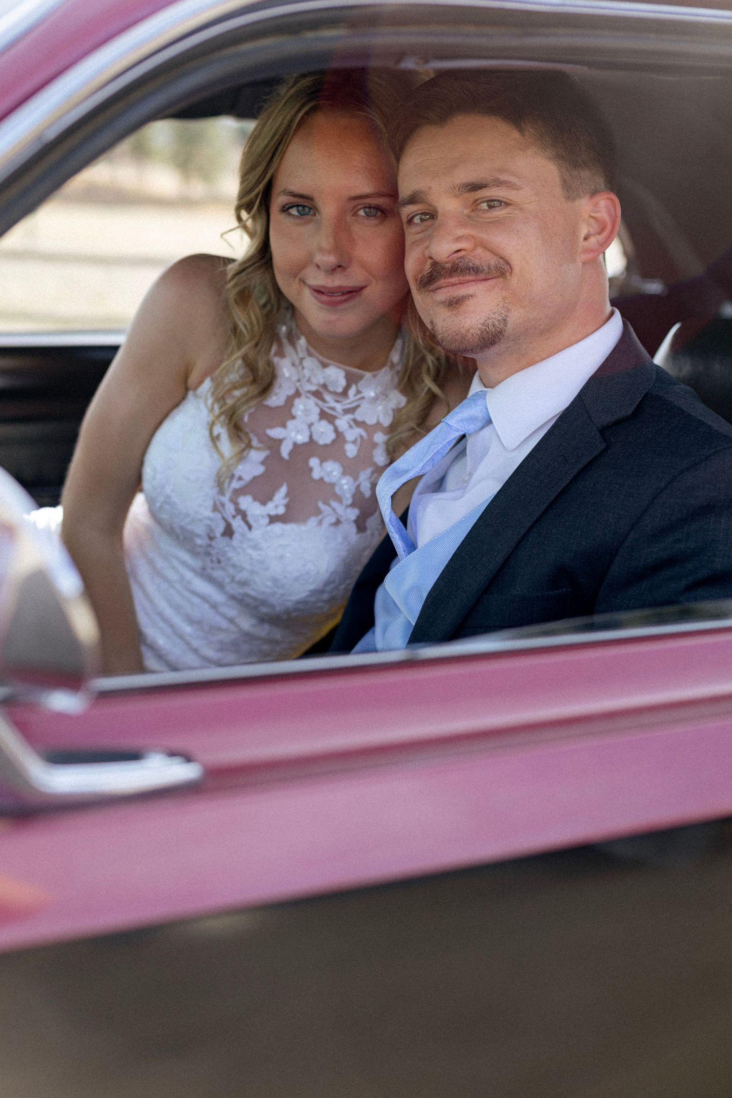 Bride and groom smile together through the window of a classic pink vintage car on their wedding day.