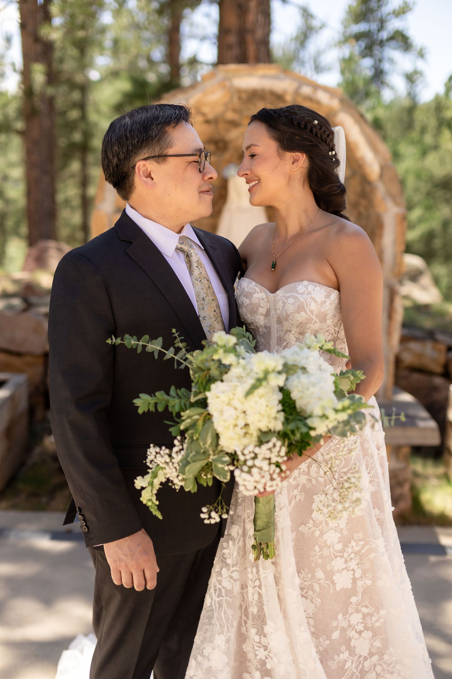A smiling bride and groom share a tender moment outdoors, bride holding a white floral bouquet.