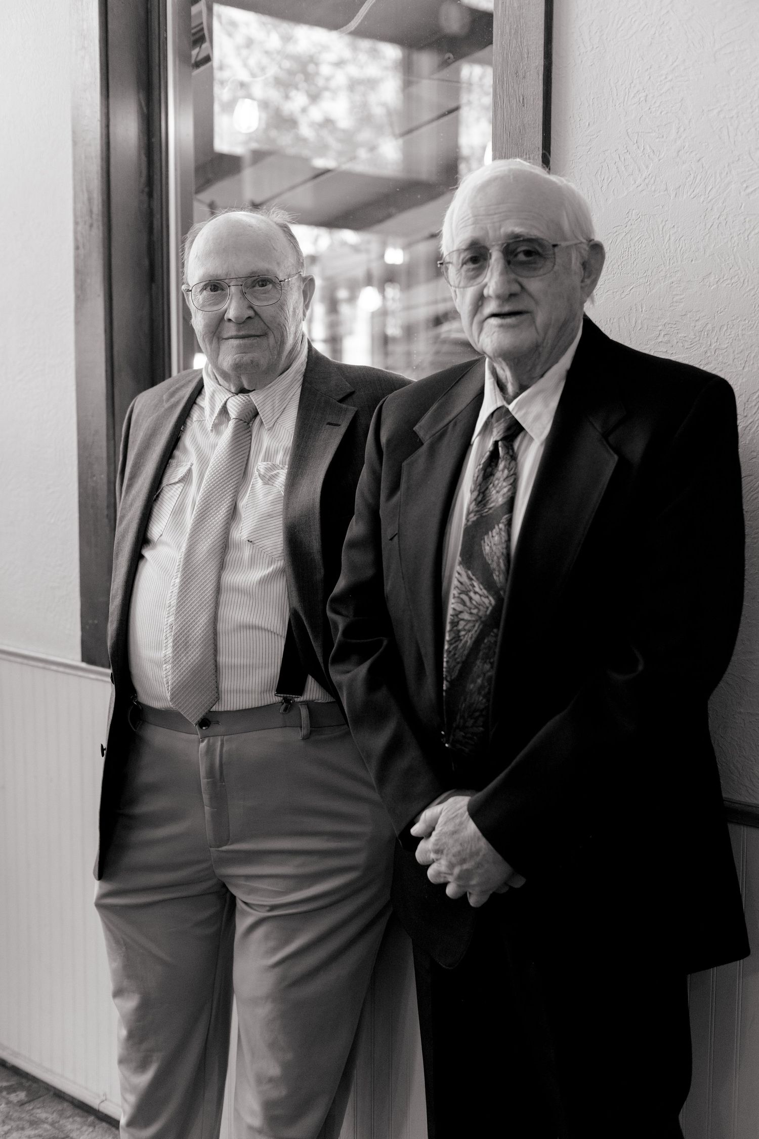 Two elderly men in formal suits smiling together in black and white portrait at wedding.
