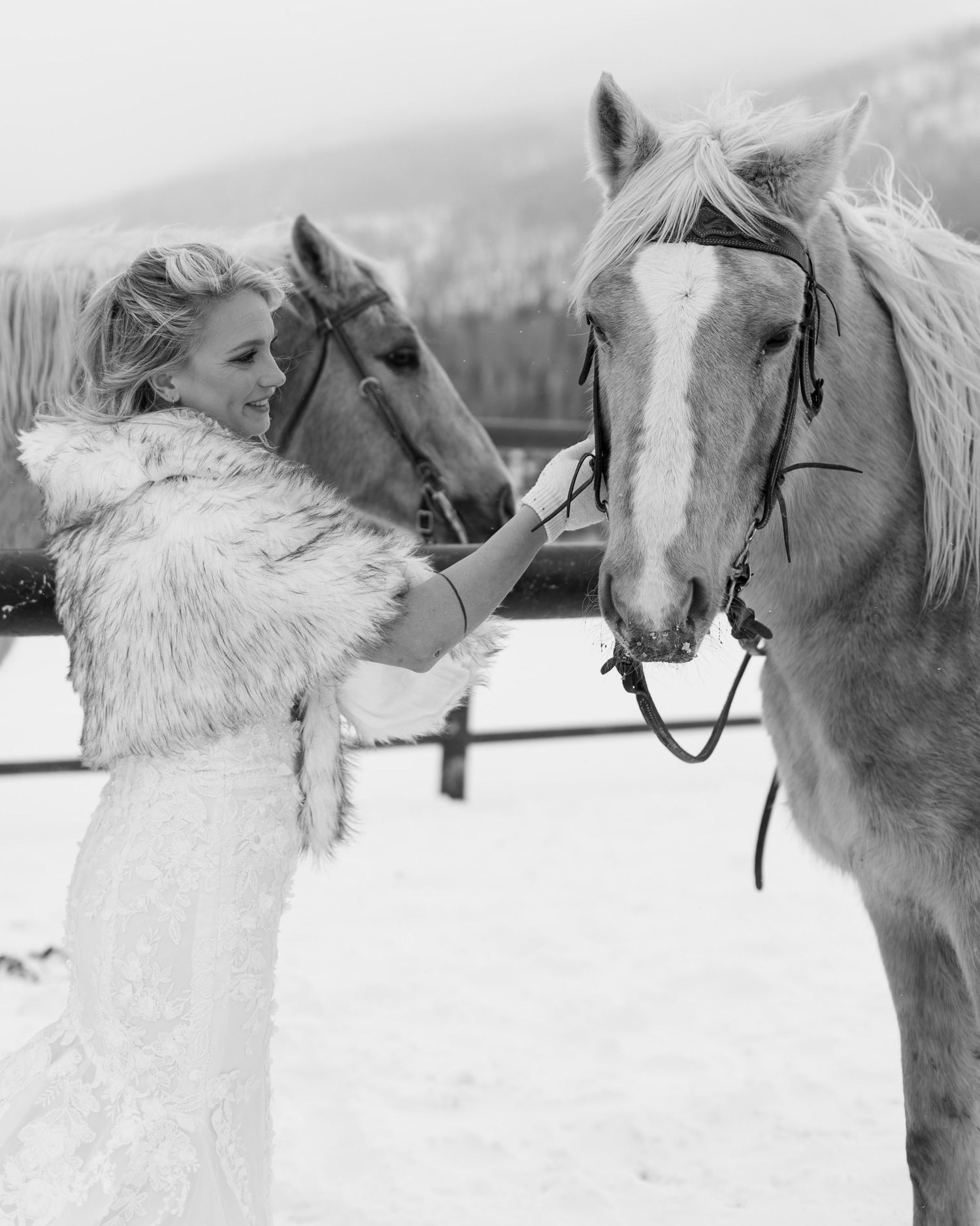 A bride in a white fur stole pets two horses during a black and white winter wedding photoshoot.