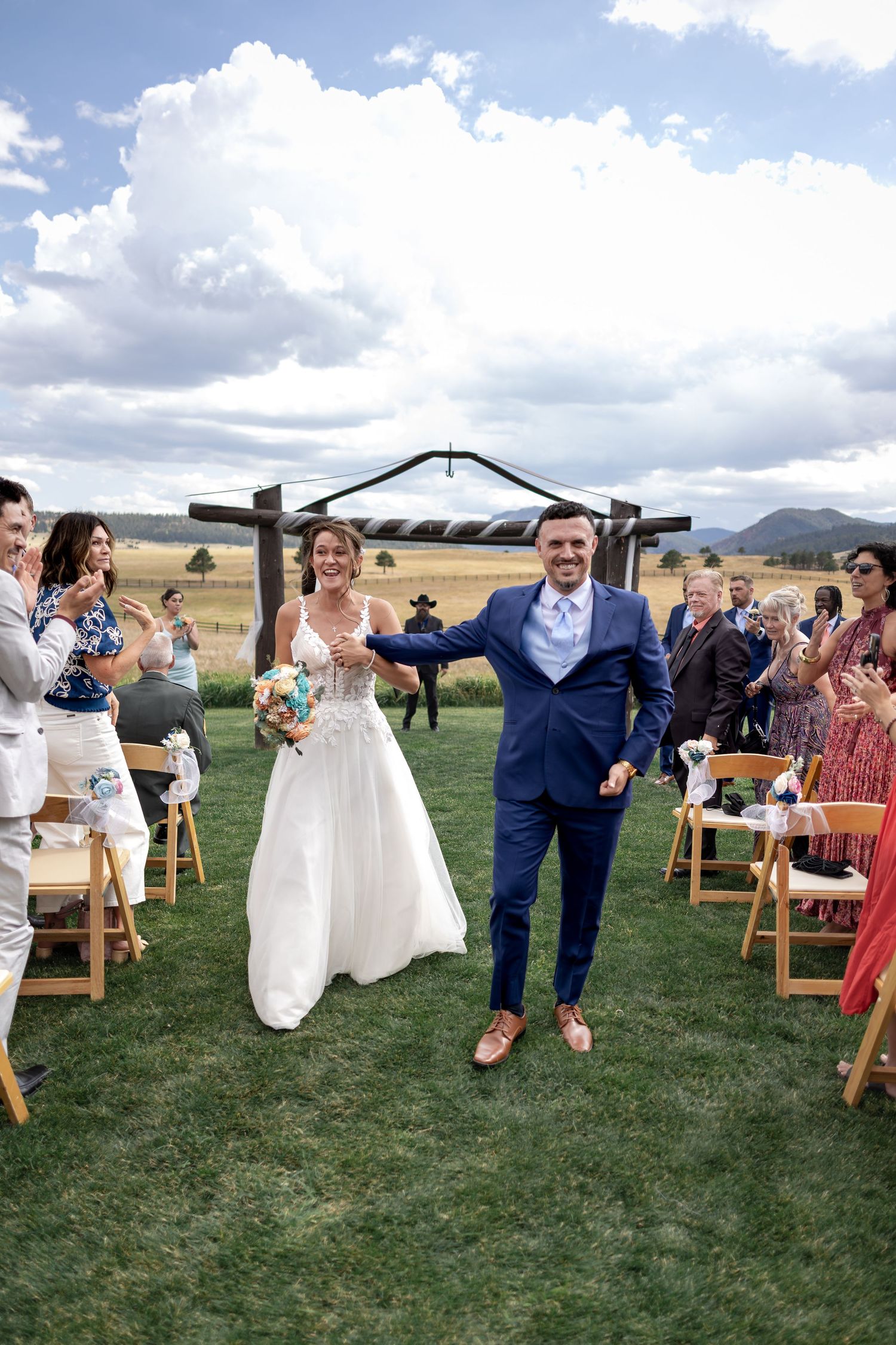 Joyful bride and groom walk down outdoor wedding aisle holding hands, guests applauding, rustic arch and mountain backdrop.