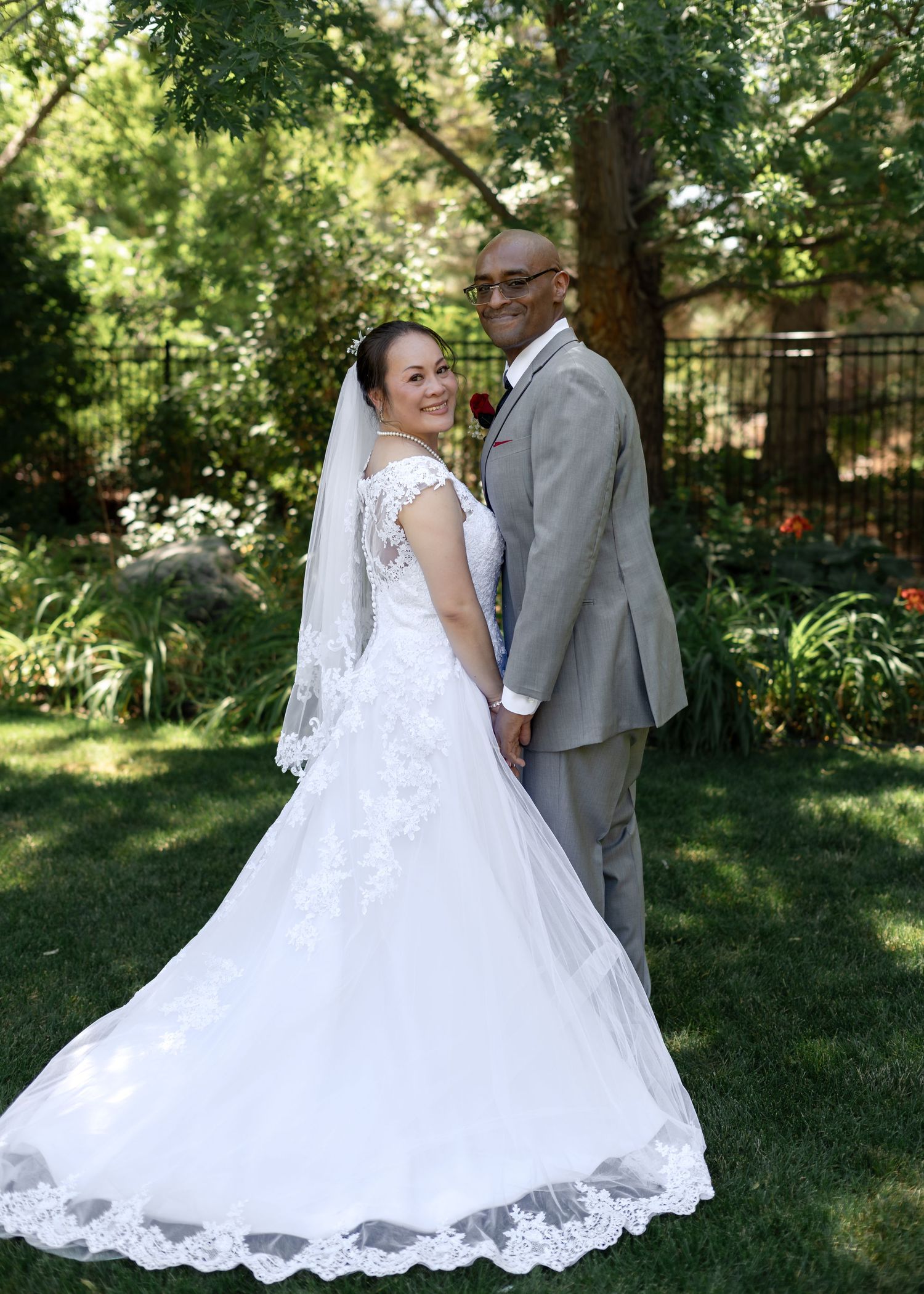 Interracial bride and groom posing back-to-back in lace wedding gown and grey suit in garden.