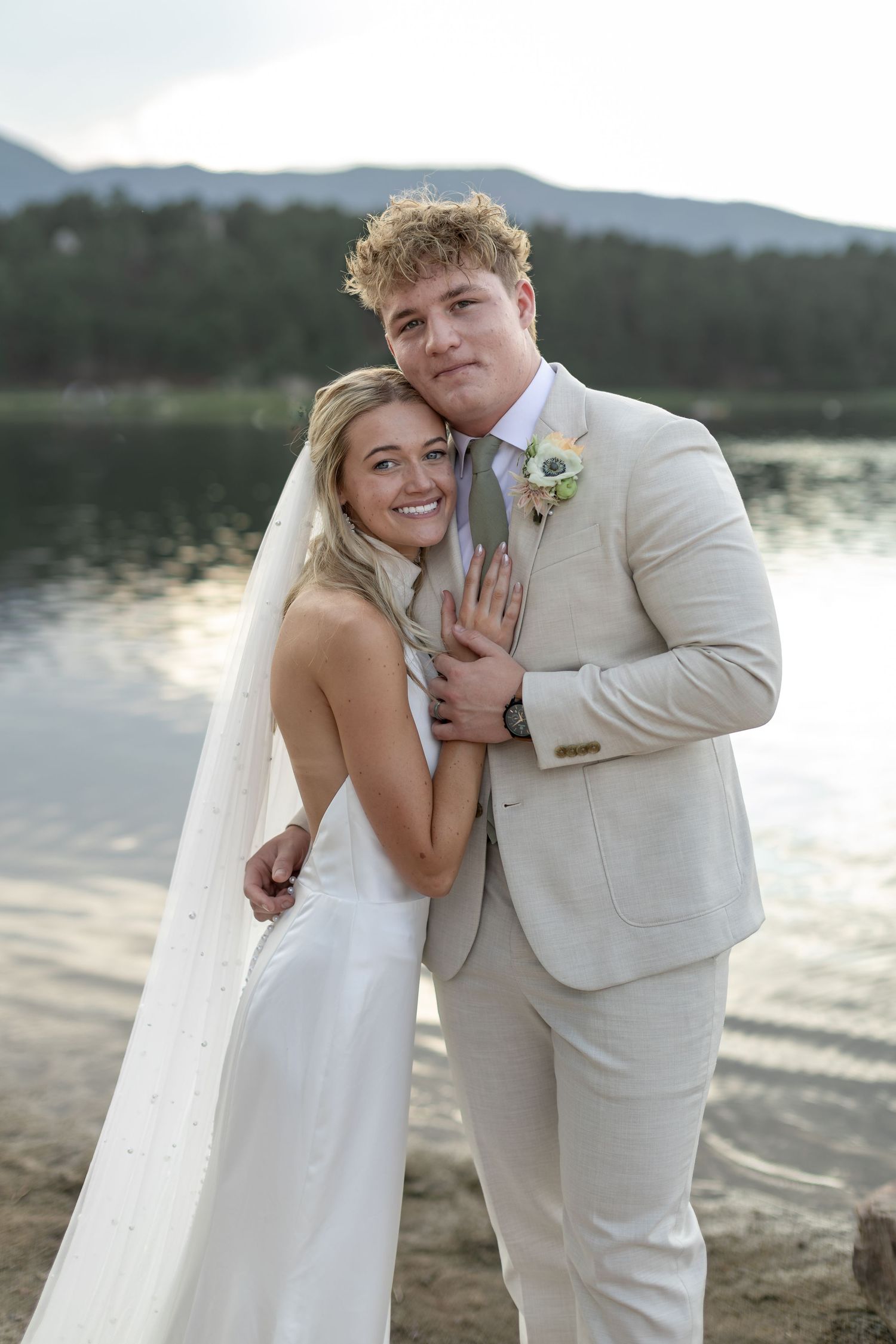 Bride and groom posing by a serene lake at sunset, she in a white gown with veil, he in a beige suit.