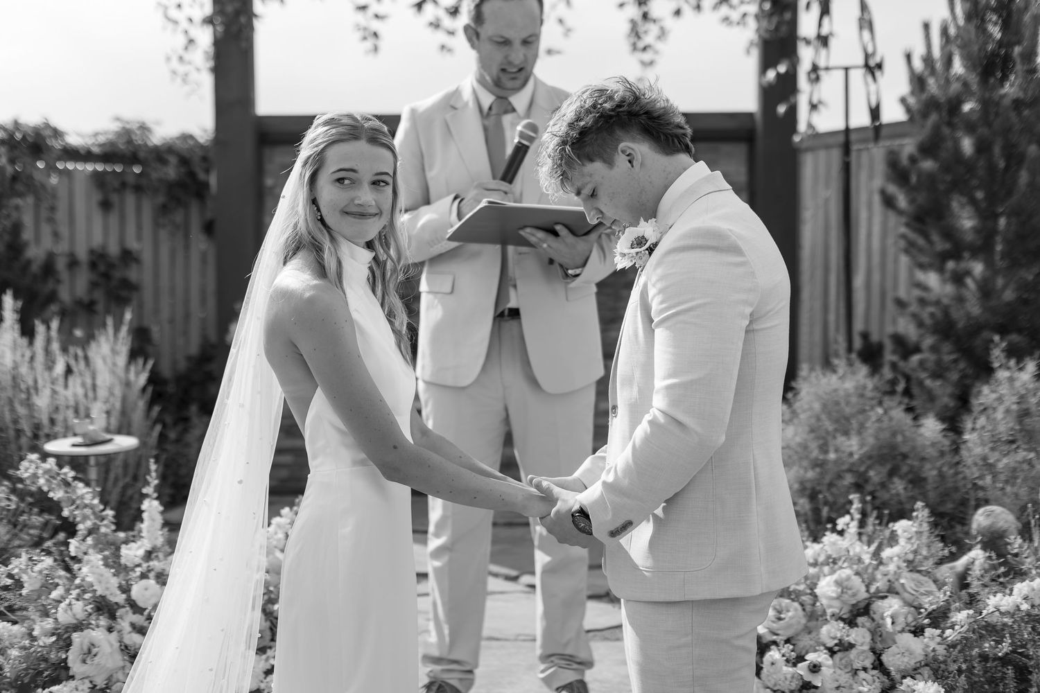 Black and white photo of bride and groom holding hands at outdoor wedding ceremony with officiant.