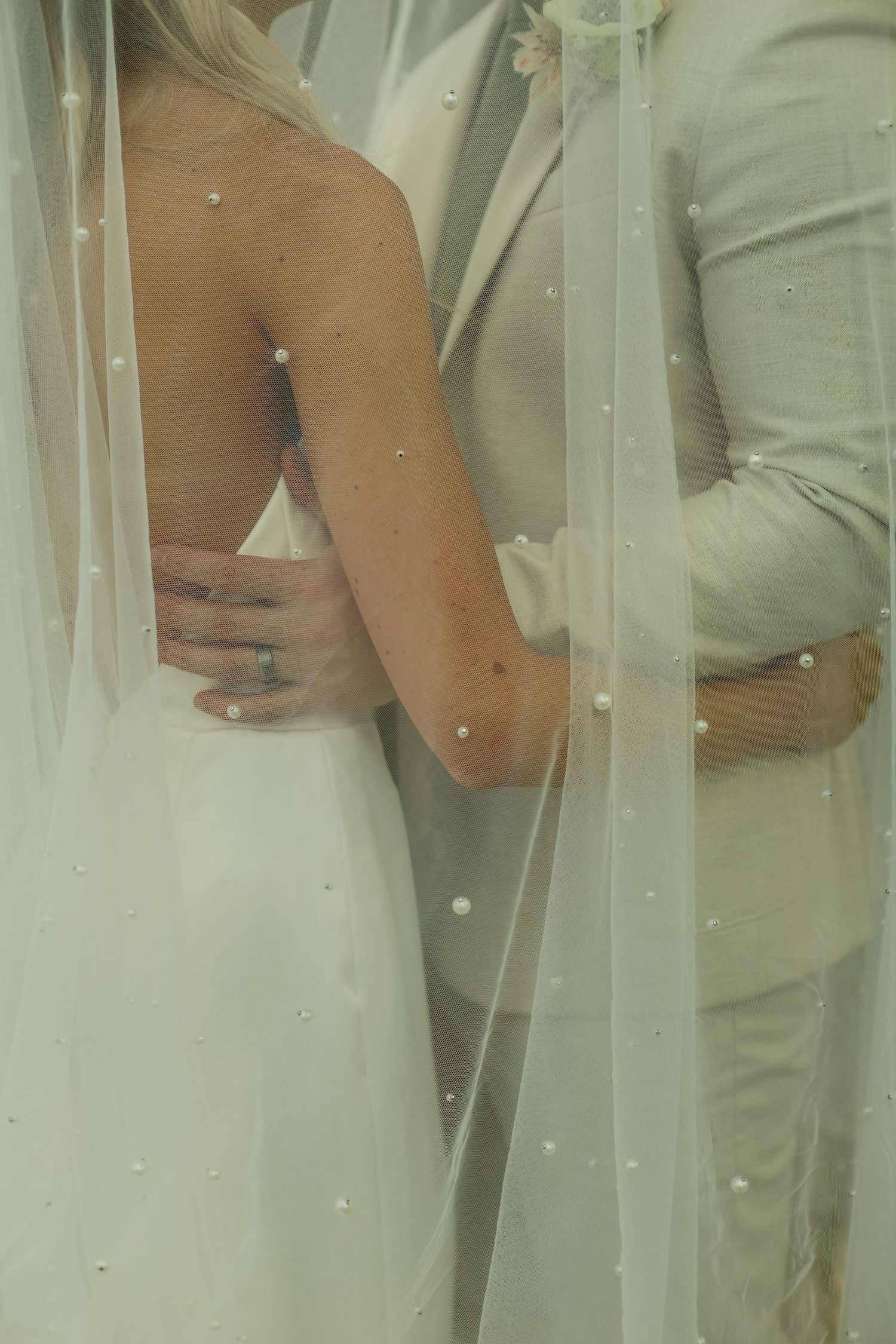Bride and groom embracing, seen through a pearl-embellished white wedding veil, wearing elegant white and cream attire.